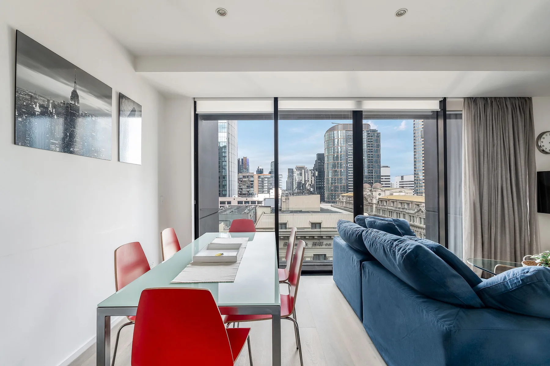 A dining area with a table and red seating overlooks city buildings through large windows.