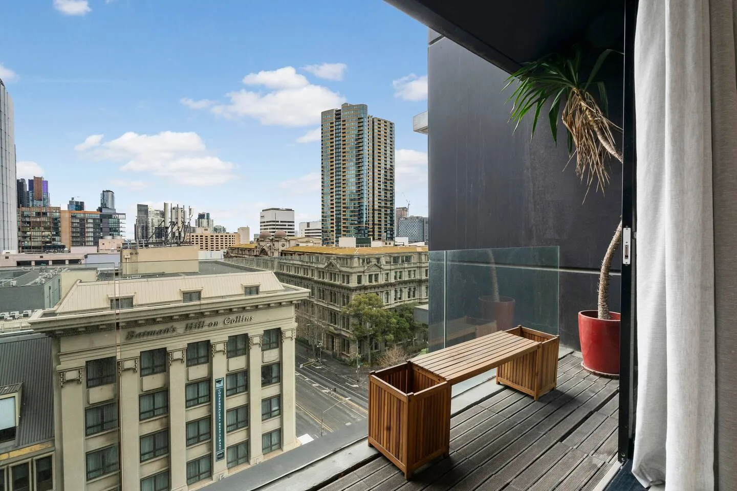 A balcony overlooks city buildings, with wooden seating and a potted plant.