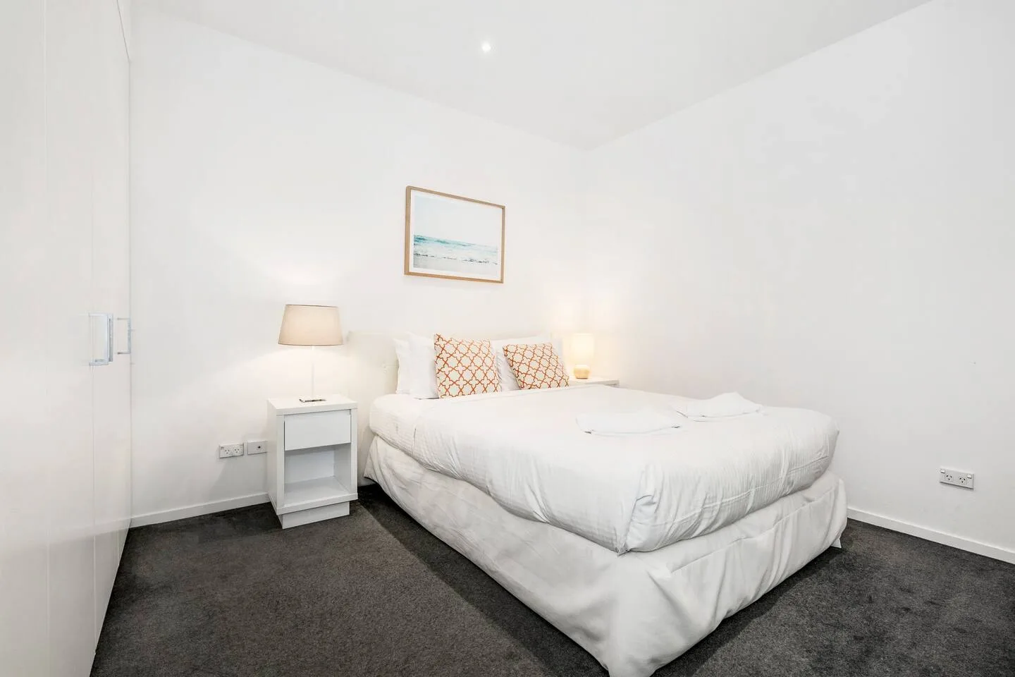 A bright bedroom has a neatly made bed with patterned cushions and a bedside table.