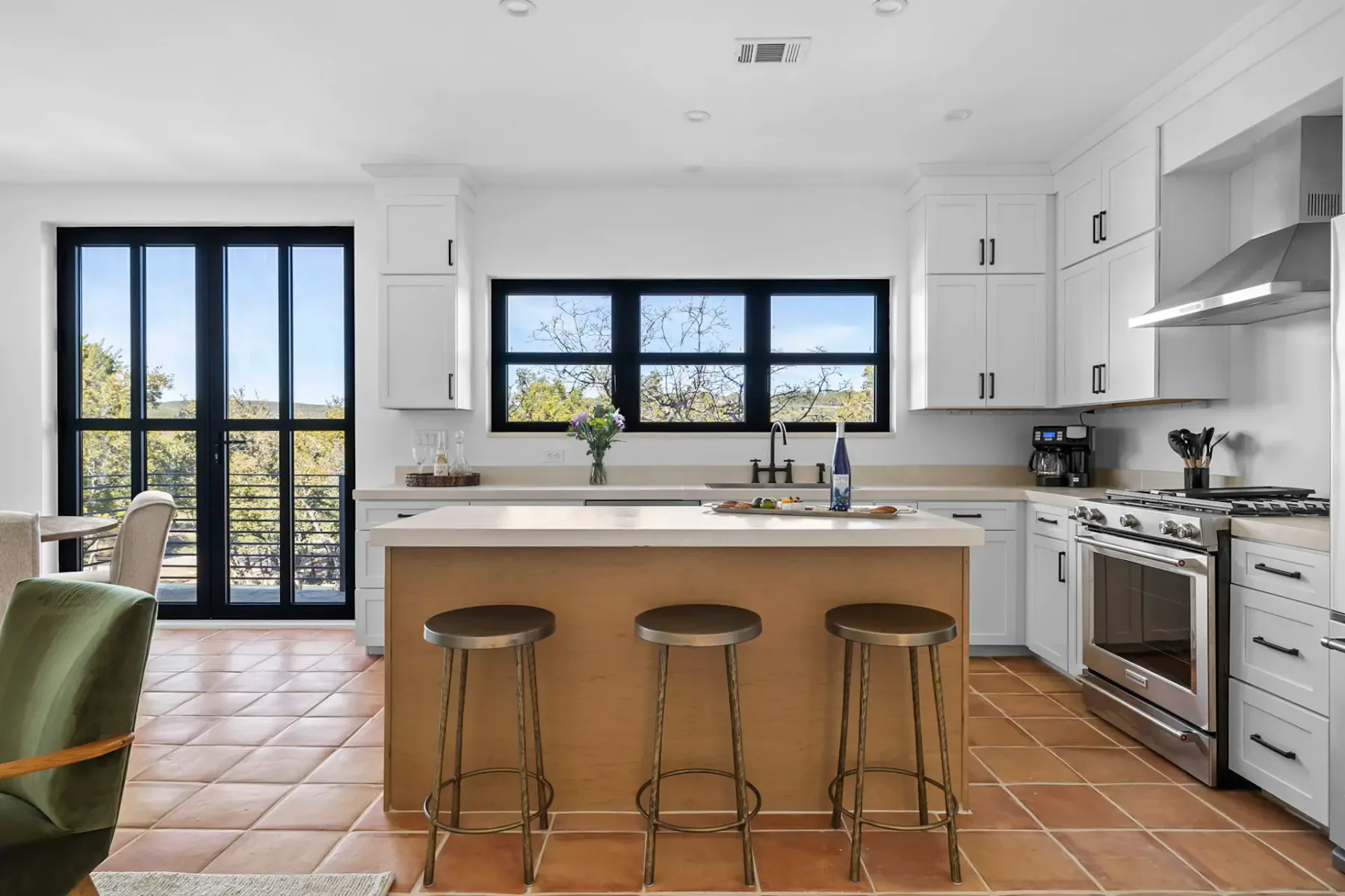 Modern kitchen with white cabinets, stainless steel appliances, central island with wooden stools, and large windows providing natural light.
