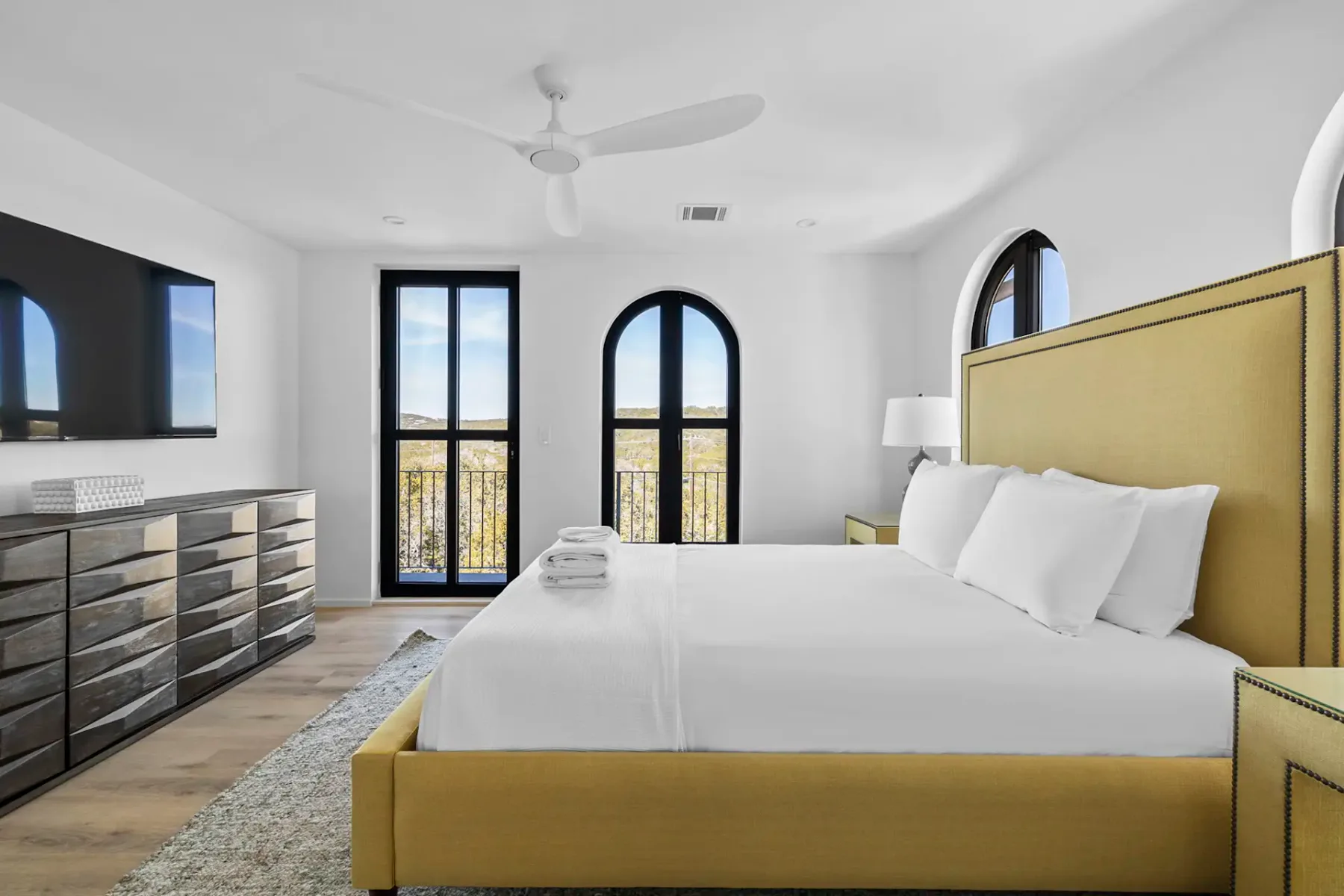 Modern bedroom with a yellow bed frame, arched black-framed windows, round mirrors, and a soft rug on wooden floors, creating a bright, airy feel.