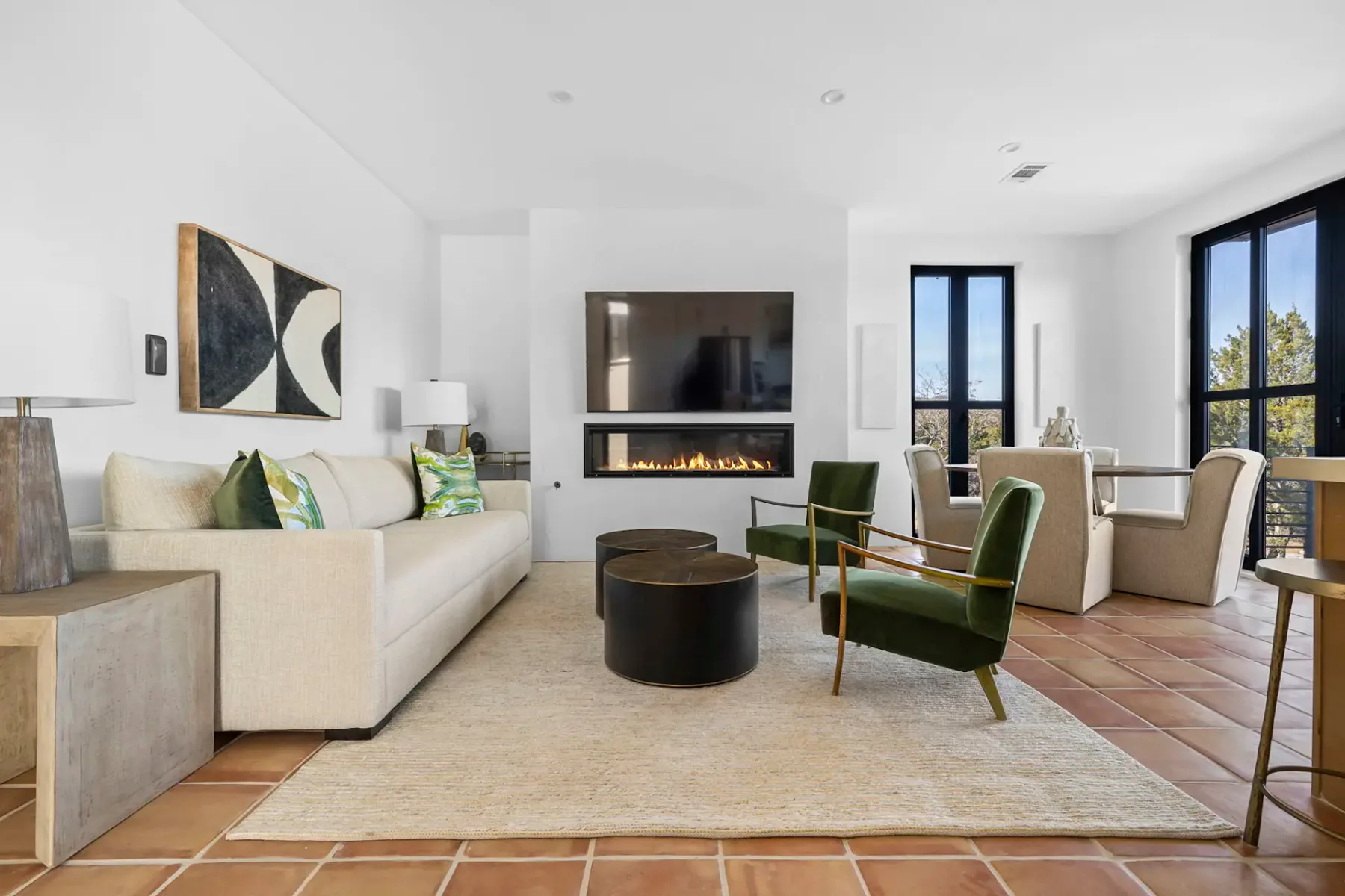 Modern living room with a beige sofa, abstract art, fireplace, green chairs, round coffee tables, and large windows with natural light.