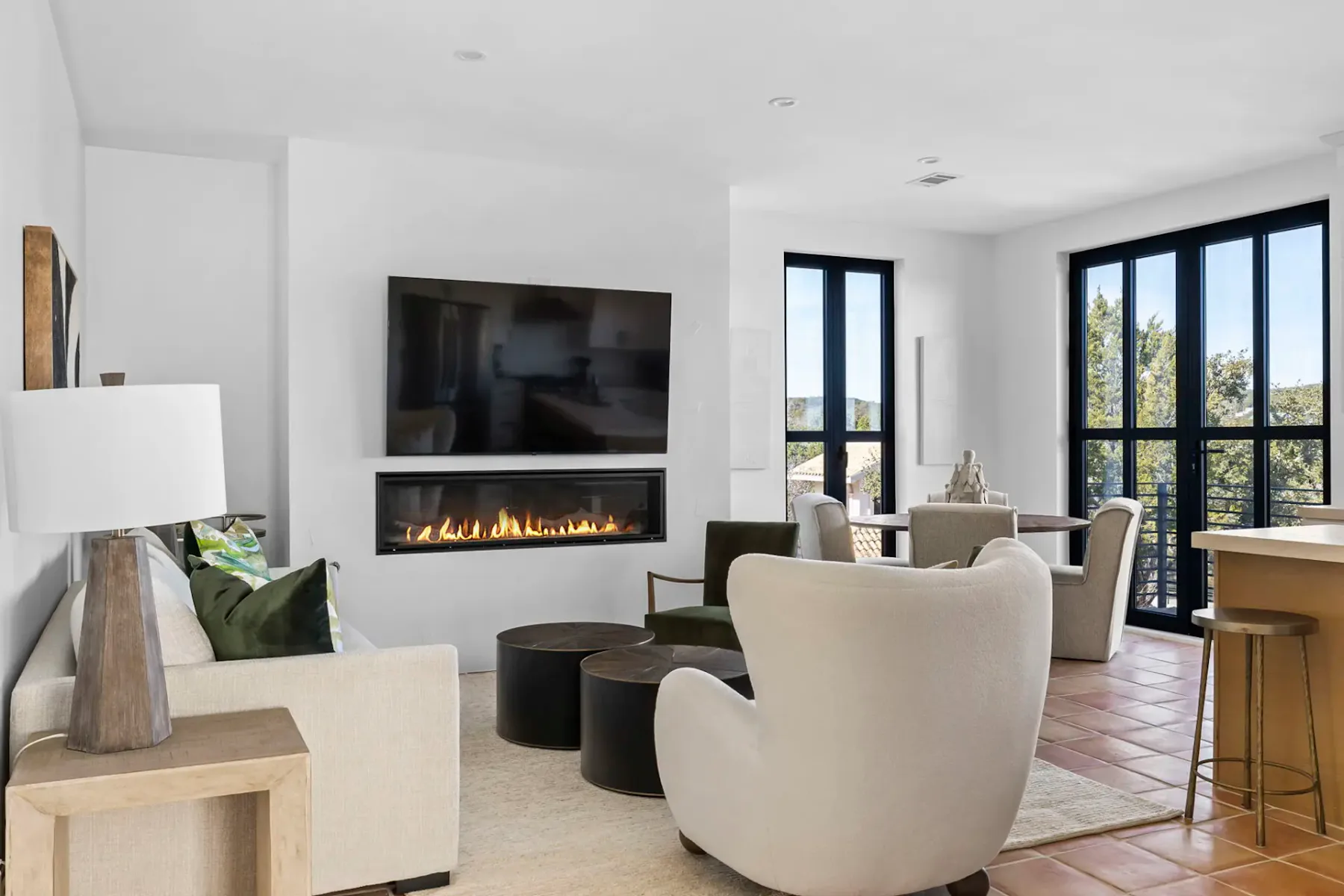 Modern living room with a beige sofa, abstract art, fireplace, green chairs, round coffee tables, and large windows with natural light.