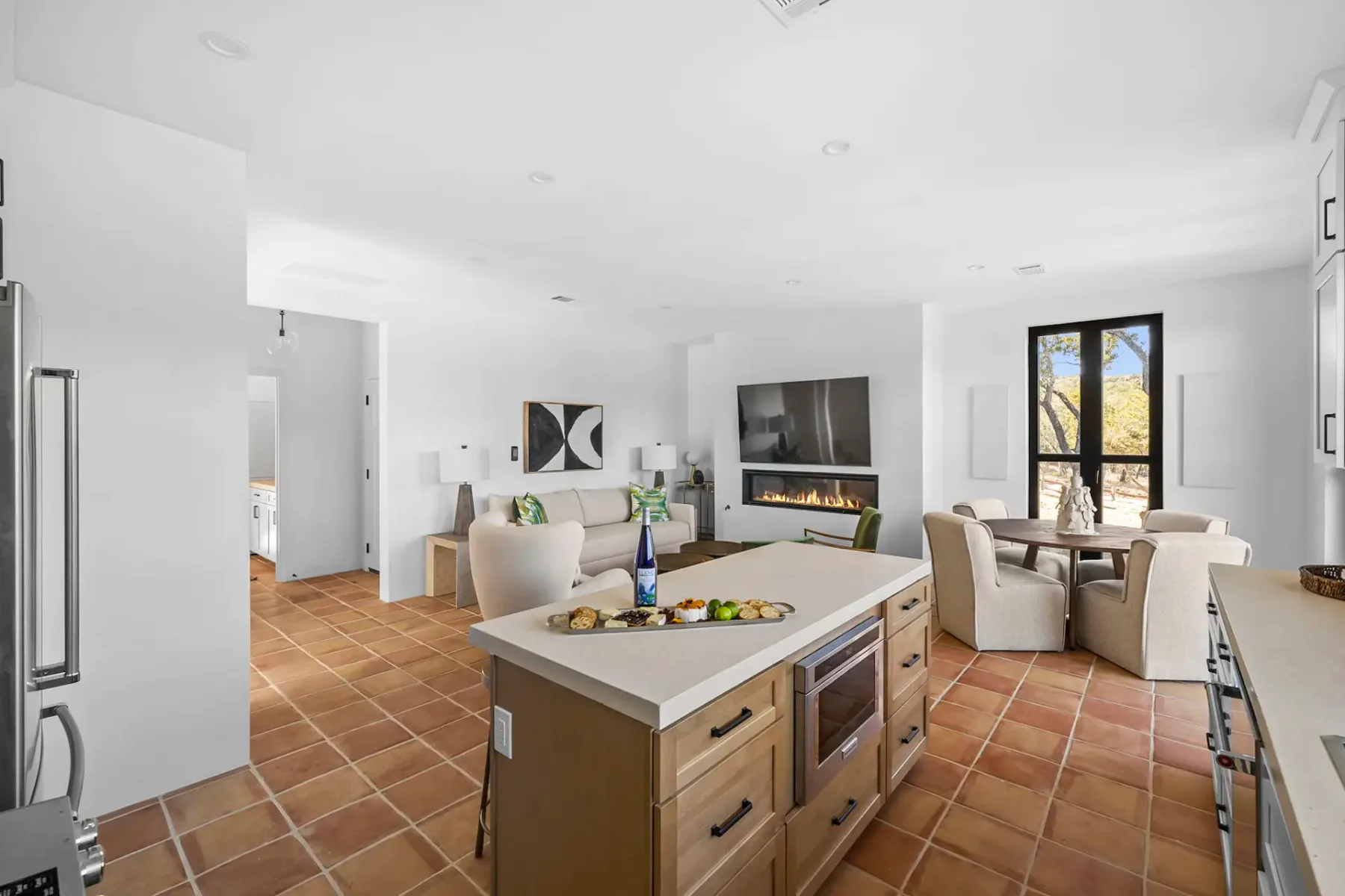 Bright kitchen with terracotta tile floor, island counter, modern cabinets, round dining table, large windows, and a wall-mounted TV with a fireplace.