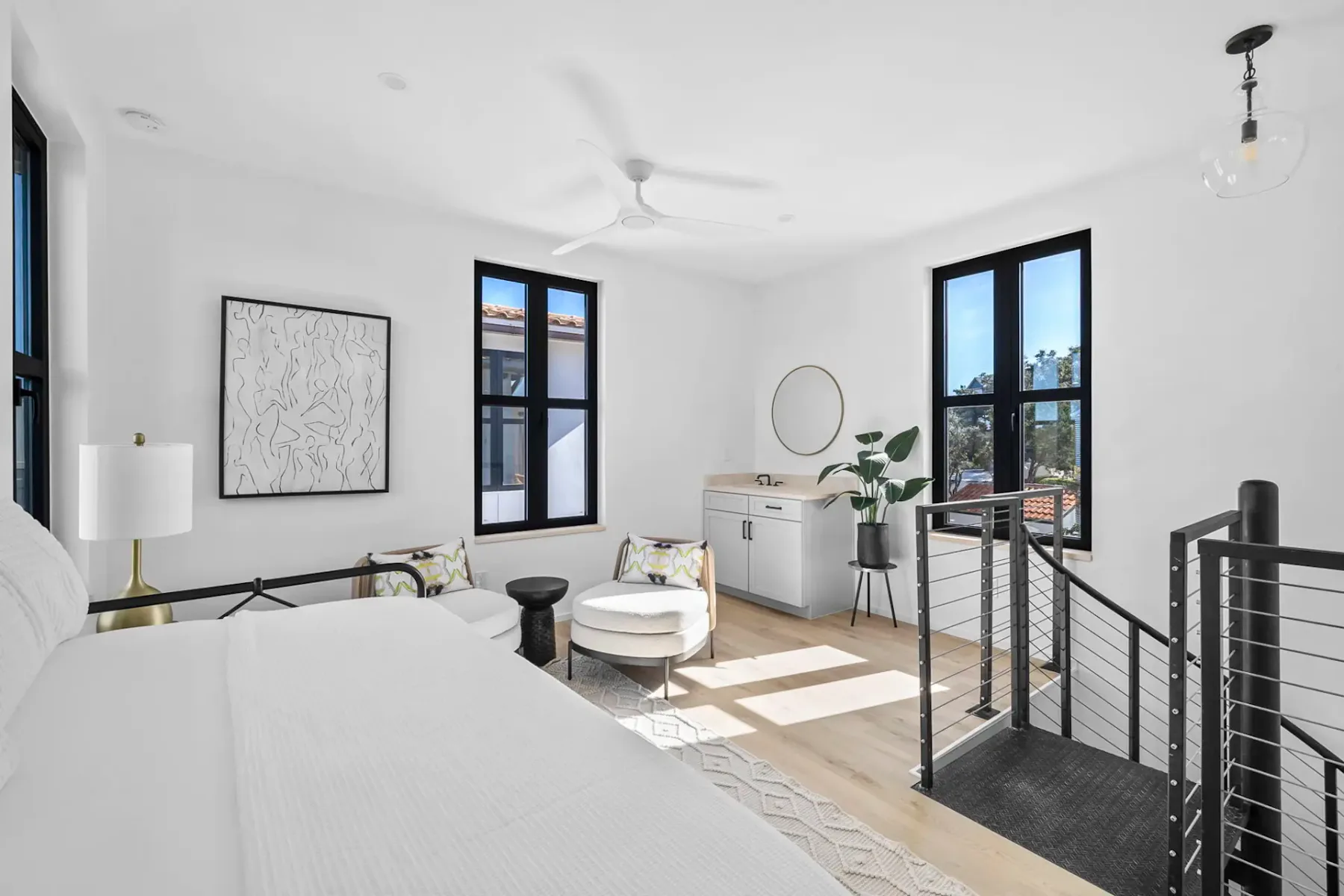 Bright, modern bedroom with large windows, white walls, sleek furniture, a wall-mounted TV, and a ceiling fan. Neutral tones and minimalist decor.