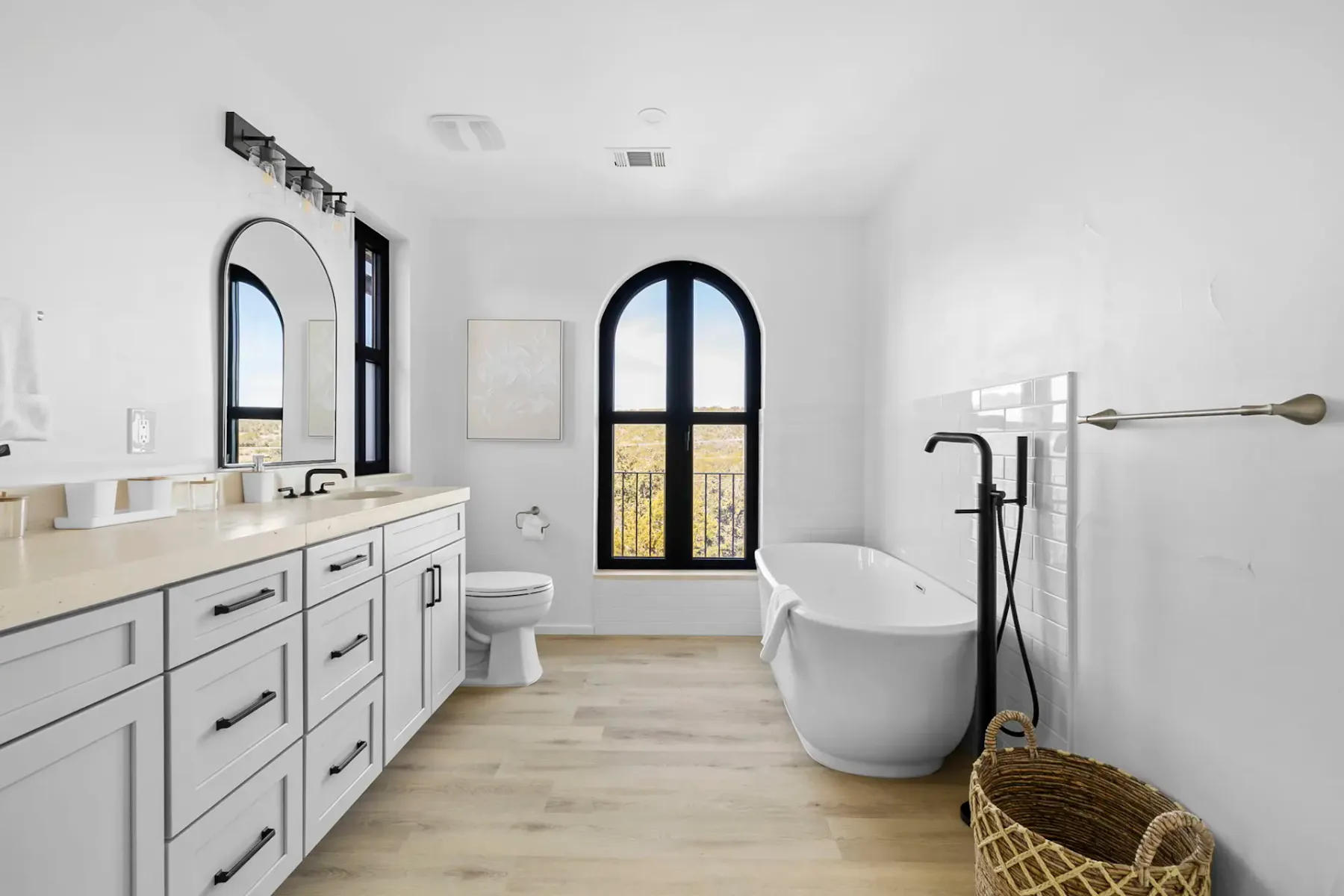 A bright bathroom has white cabinetry, a freestanding tub, and tall arched windows.