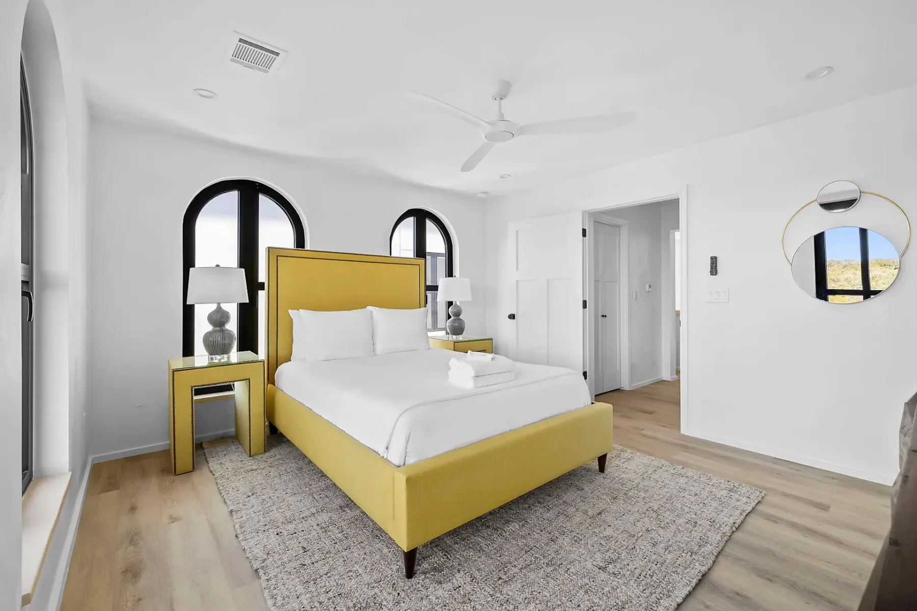 Modern bedroom with a yellow bed frame, arched black-framed windows, round mirrors, and a soft rug on wooden floors, creating a bright, airy feel.