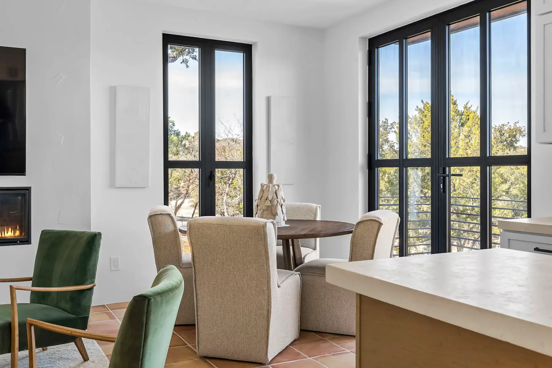 A modern dining area with a round table, beige chairs, and large windows overlooking a green landscape. A fireplace is visible on the left.