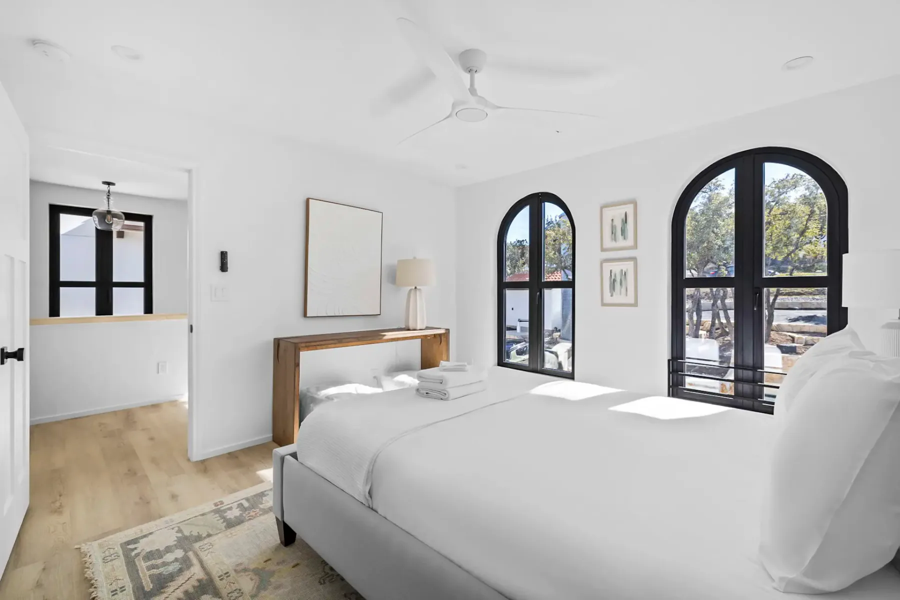 Bright bedroom with white walls, a large bed, wooden console table, two stools, arched windows, and a ceiling fan. Minimalist decor.