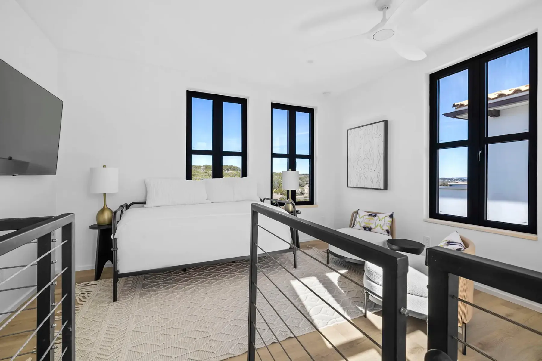 Bright, modern bedroom with large windows, white walls, sleek furniture, a wall-mounted TV, and a ceiling fan. Neutral tones and minimalist decor.