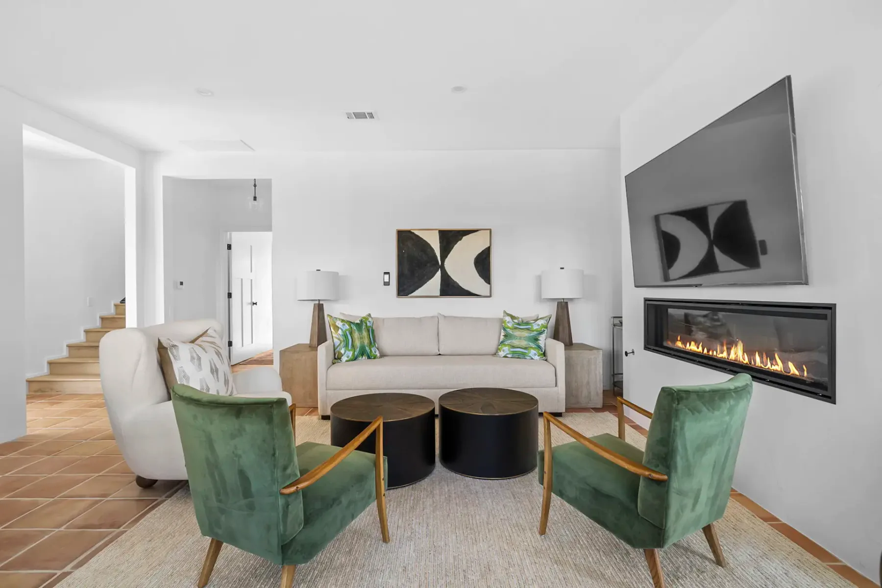 Modern living room with a beige sofa, abstract art, fireplace, green chairs, round coffee tables, and large windows with natural light.