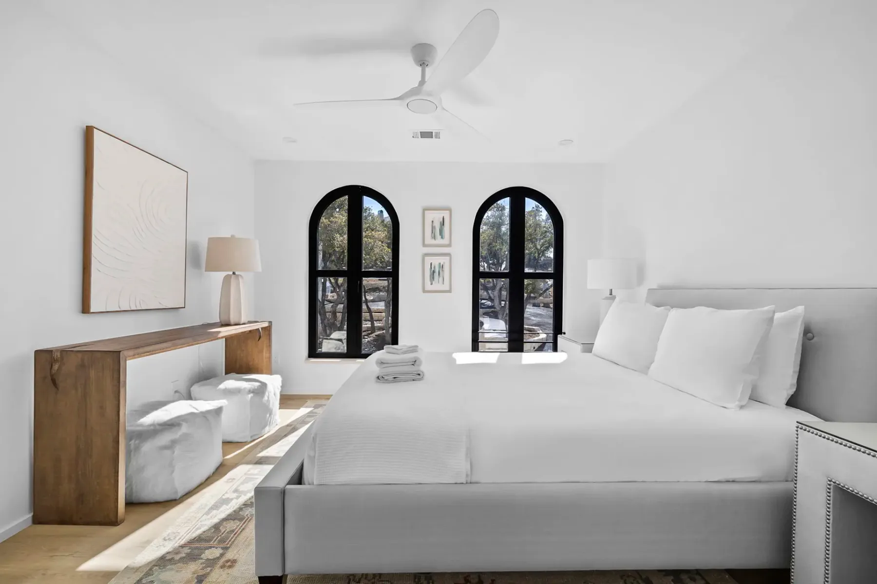 Bright bedroom with white walls, a large bed, wooden console table, two stools, arched windows, and a ceiling fan. Minimalist decor.