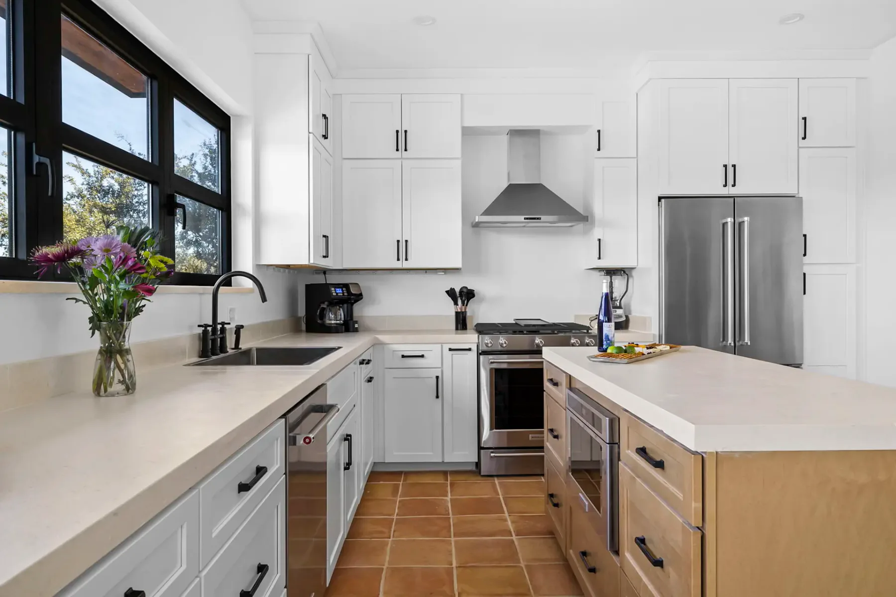 Modern kitchen with white cabinets, stainless steel appliances, a central island with light wood finish, and a vase of flowers by the window.