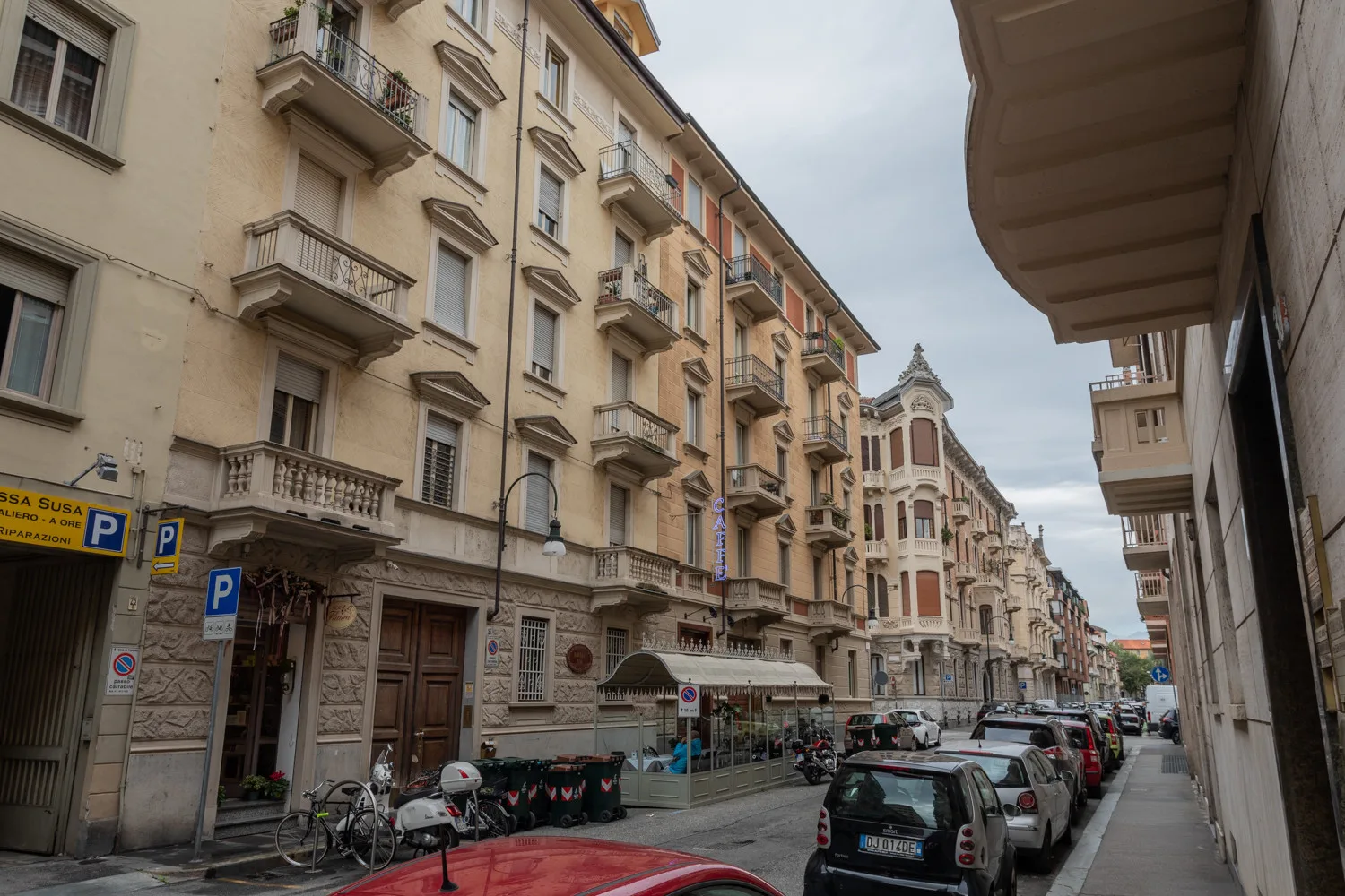 Una calle urbana flanqueada por edificios de varias plantas, balcones, coches estacionados y una acera.