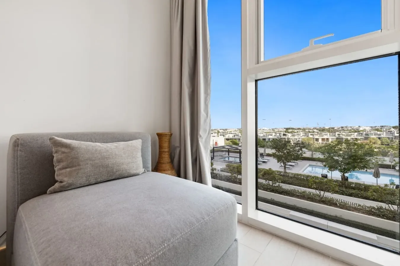 Una habitación luminosa tiene una chaise longue gris junto a una gran ventana con vistas a edificios y una piscina.
