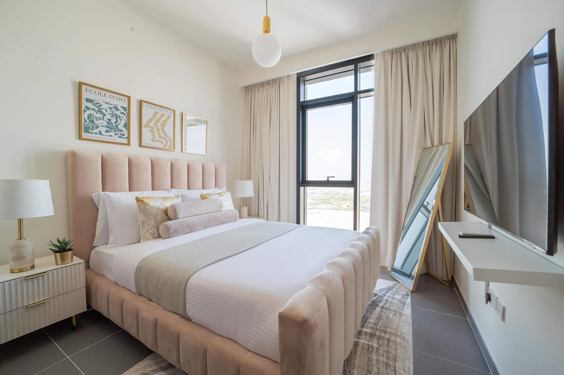 Un dormitorio luminoso muestra una cama bajo cuadros enmarcados y una ventana alta.