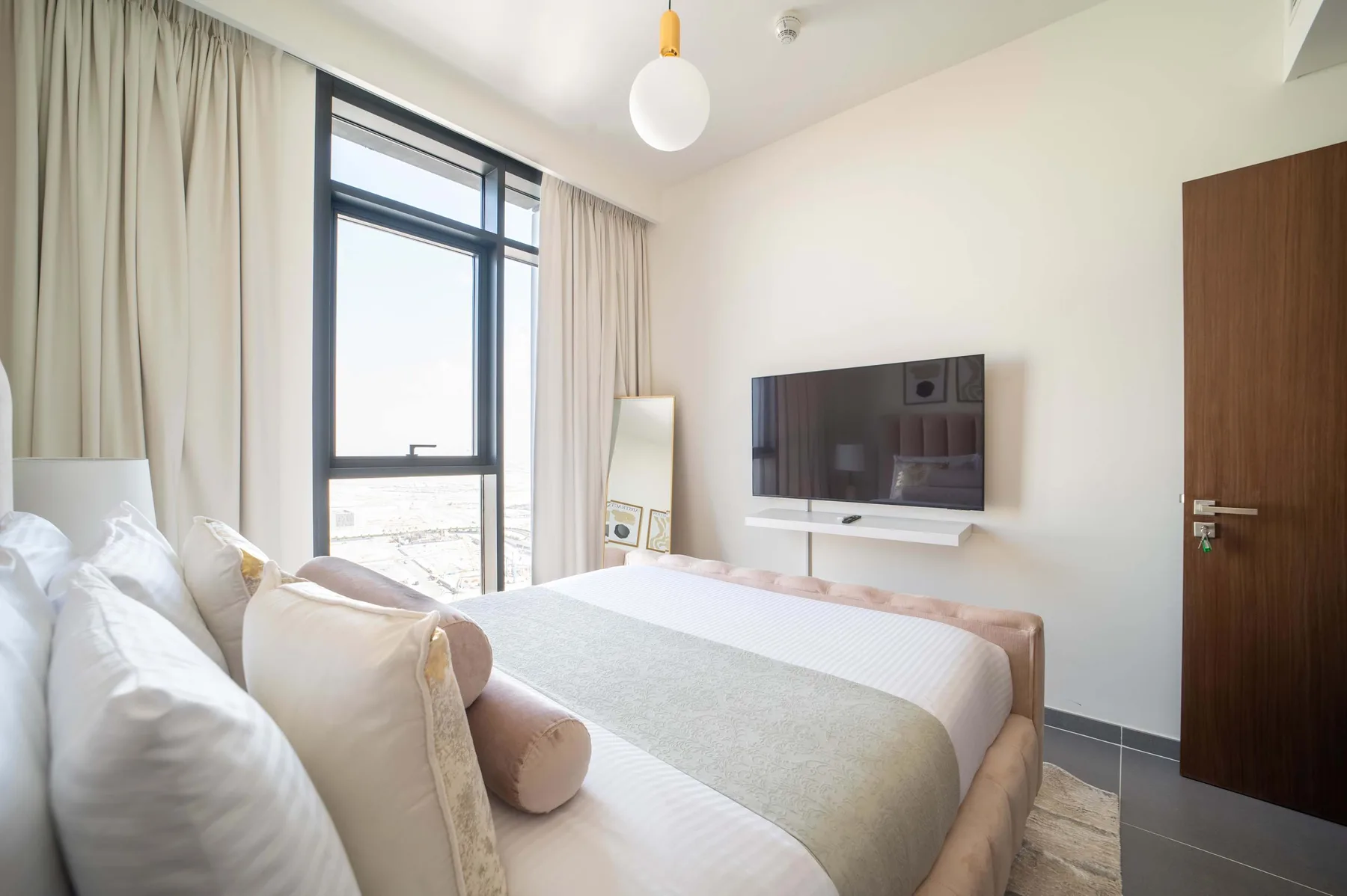 Un dormitorio luminoso con una cama junto a una ventana amplia y un televisor en la pared.