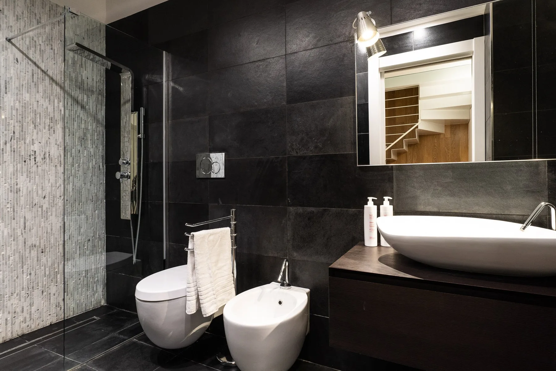 Un baño de azulejos oscuros con ducha de vidrio, inodoro, bidé y lavabo.