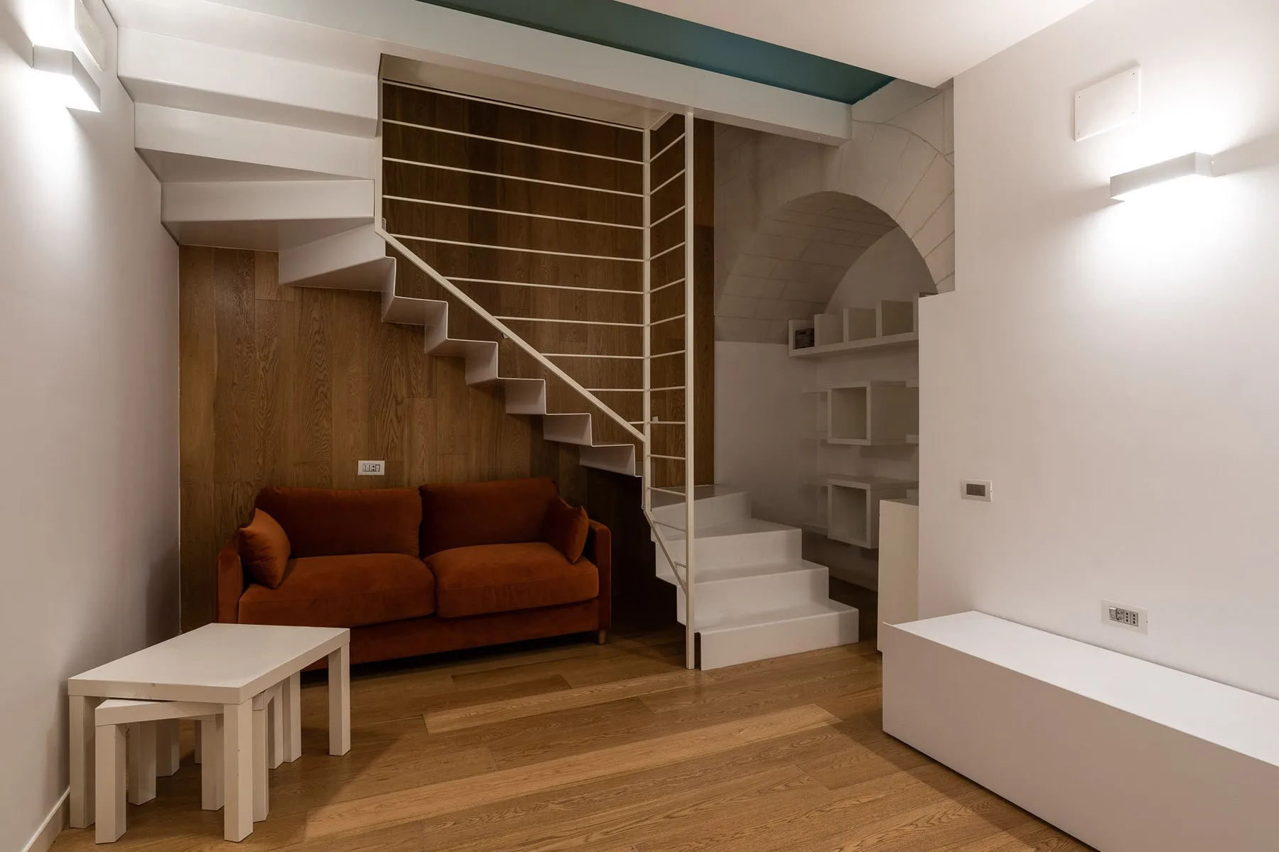 Una pequeña zona de estar con un sofá marrón bajo una escalera y estantes empotrados.