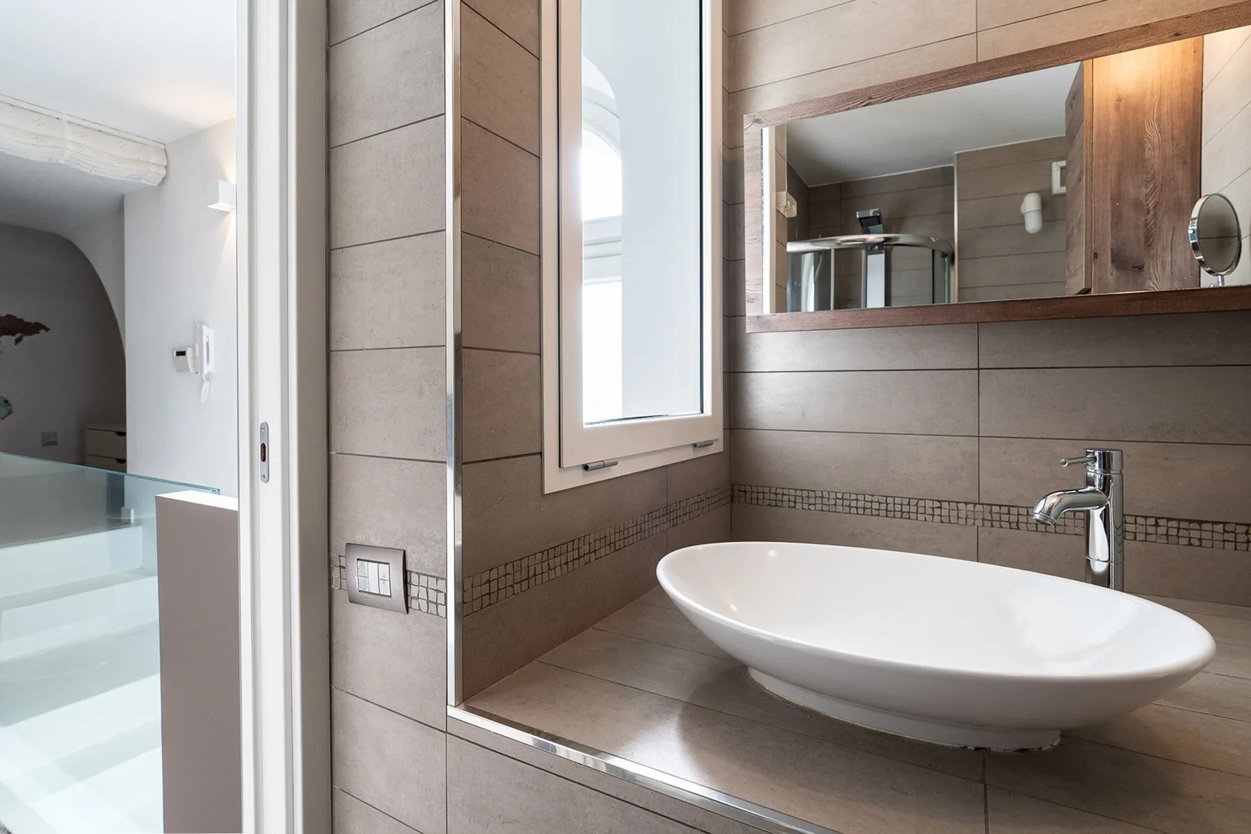 Un tocador de baño con lavabo blanco sobre encimera, espejo y una ventana cercana.