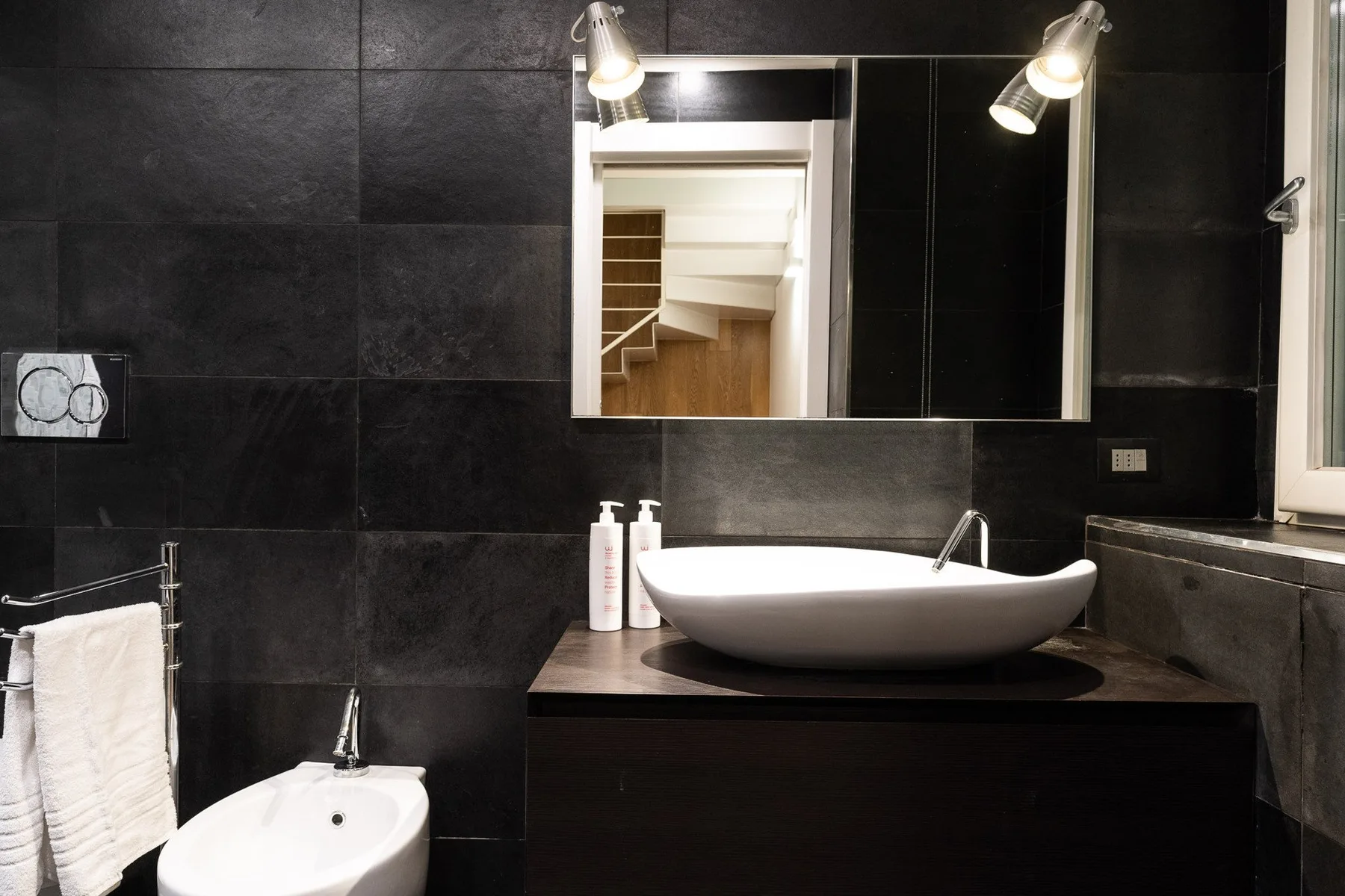 Un baño de azulejos oscuros con un lavabo sobre encimera y un espejo.