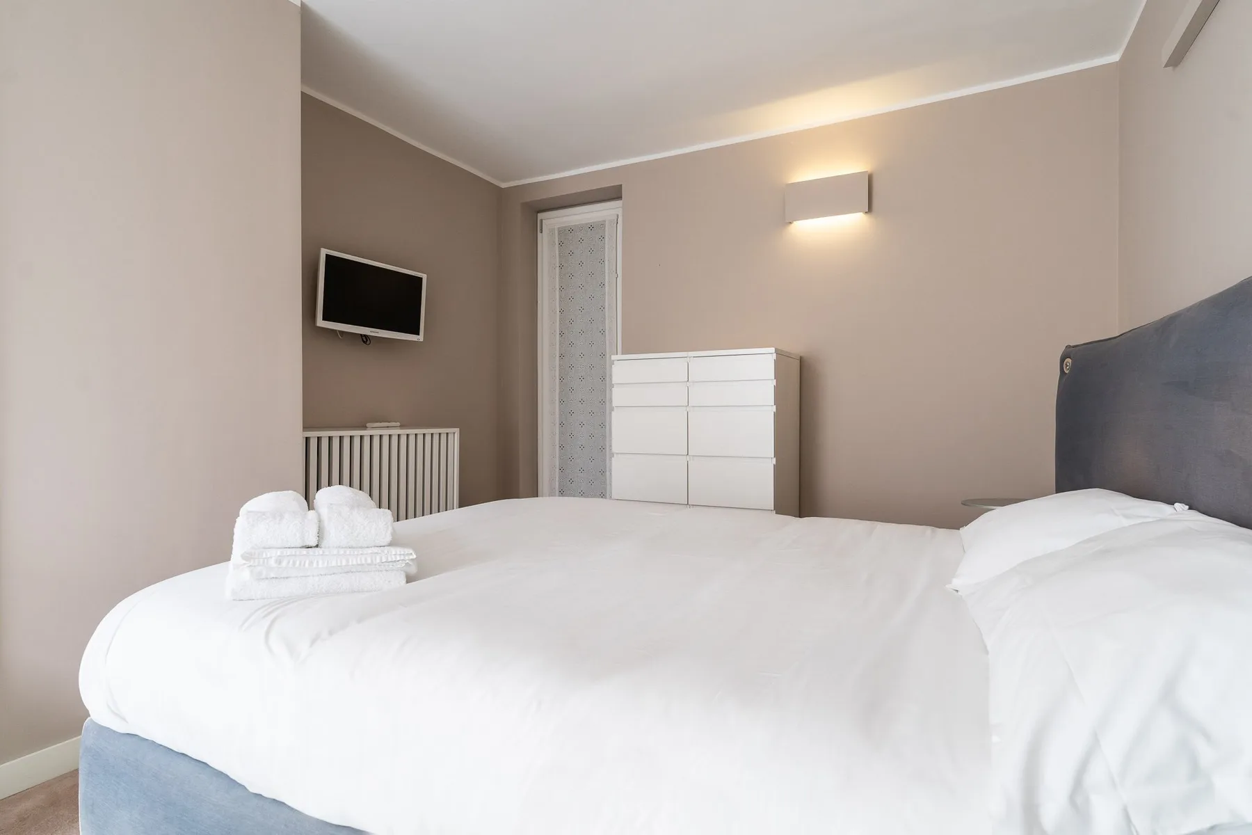 Un dormitorio con una cama blanca, una cómoda y un televisor de pared.