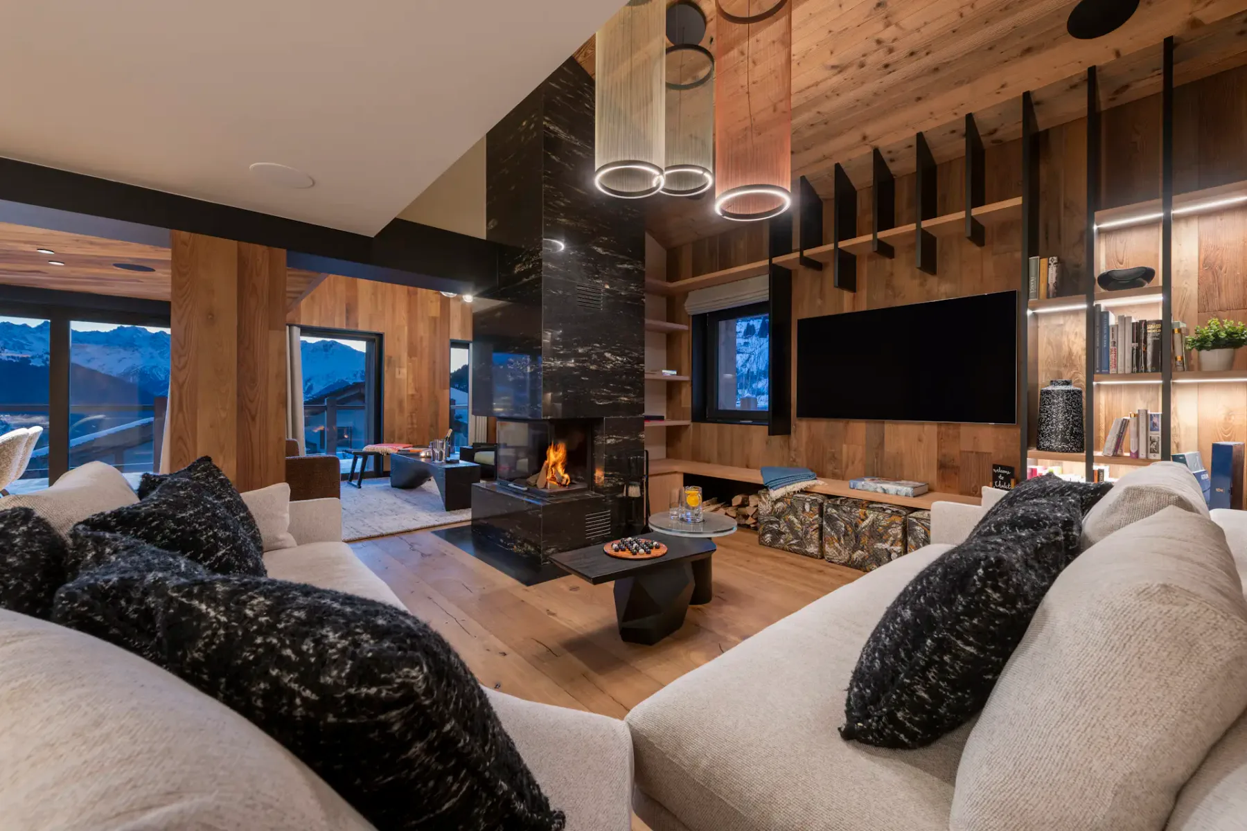 Modern living room with wooden walls and ceiling, featuring a central black fireplace, cozy sofas with dark pillows, a wall-mounted TV, and large windows with snowy mountain views.