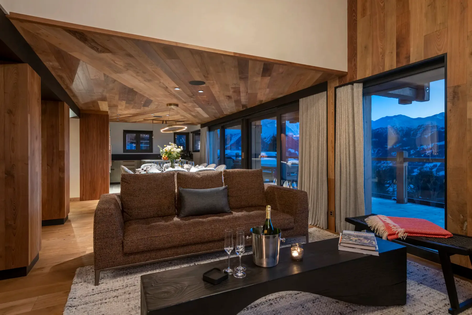 Modern living room with wooden walls and ceiling, featuring a central black fireplace, cozy sofas with dark pillows, a wall-mounted TV, and large windows with snowy mountain views.