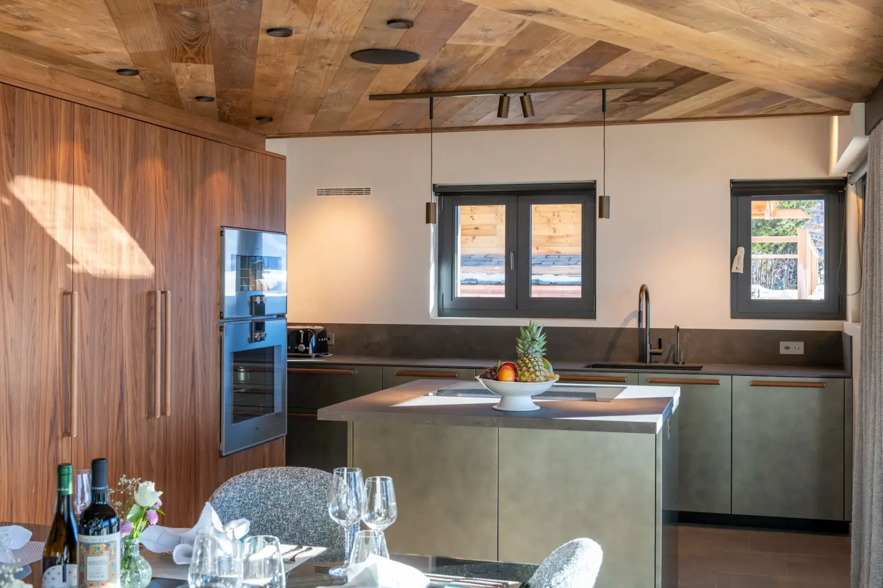 Modern kitchen with wood ceiling, wall oven, and island. Windows above sink. Dining table set with glasses, wine, and flowers in foreground.