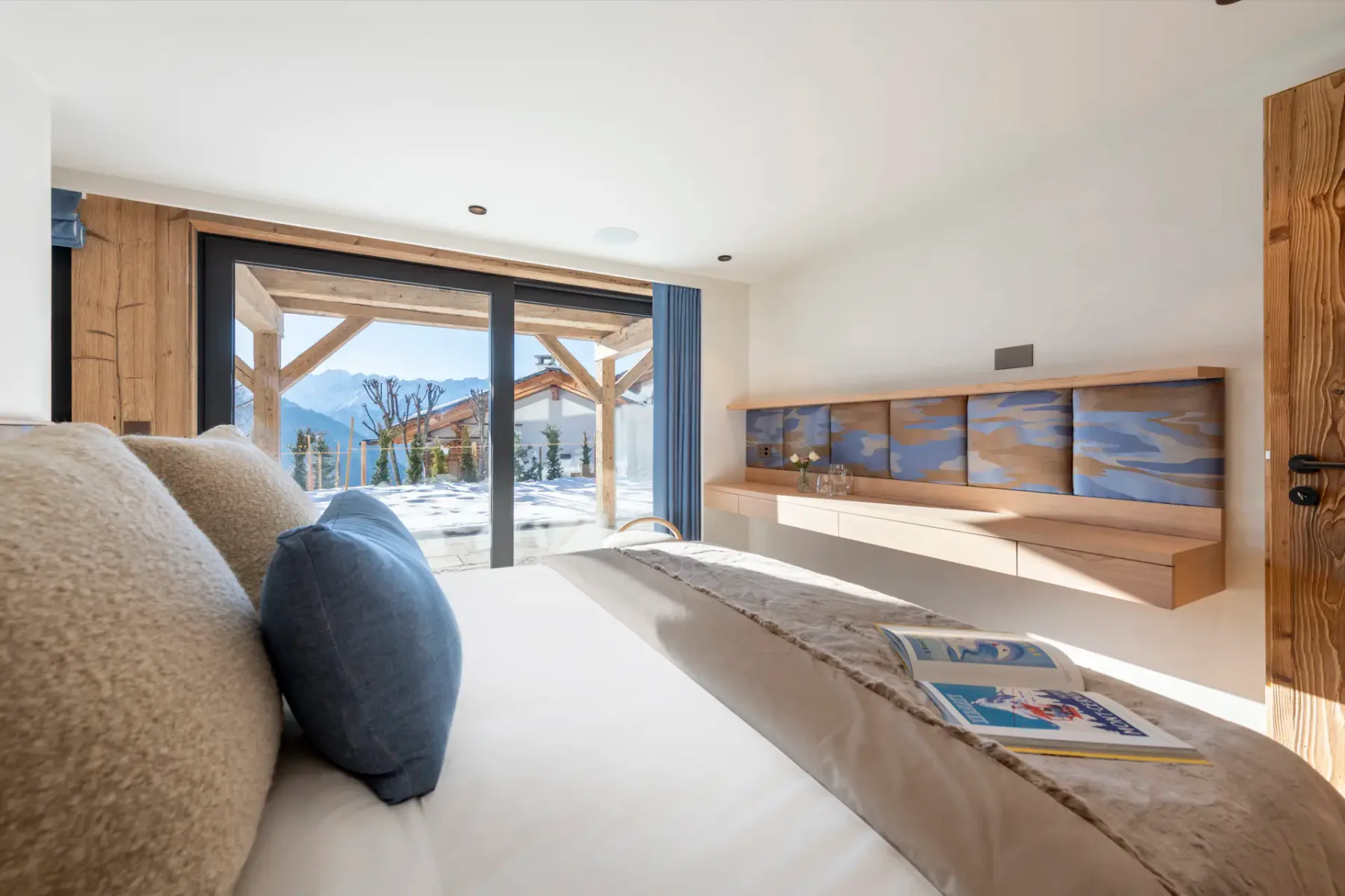 A bright bedroom with a double bed, beige and blue decor, bedside tables, and a scenic mountain view through large sliding glass doors. An open magazine rests on the bed.
