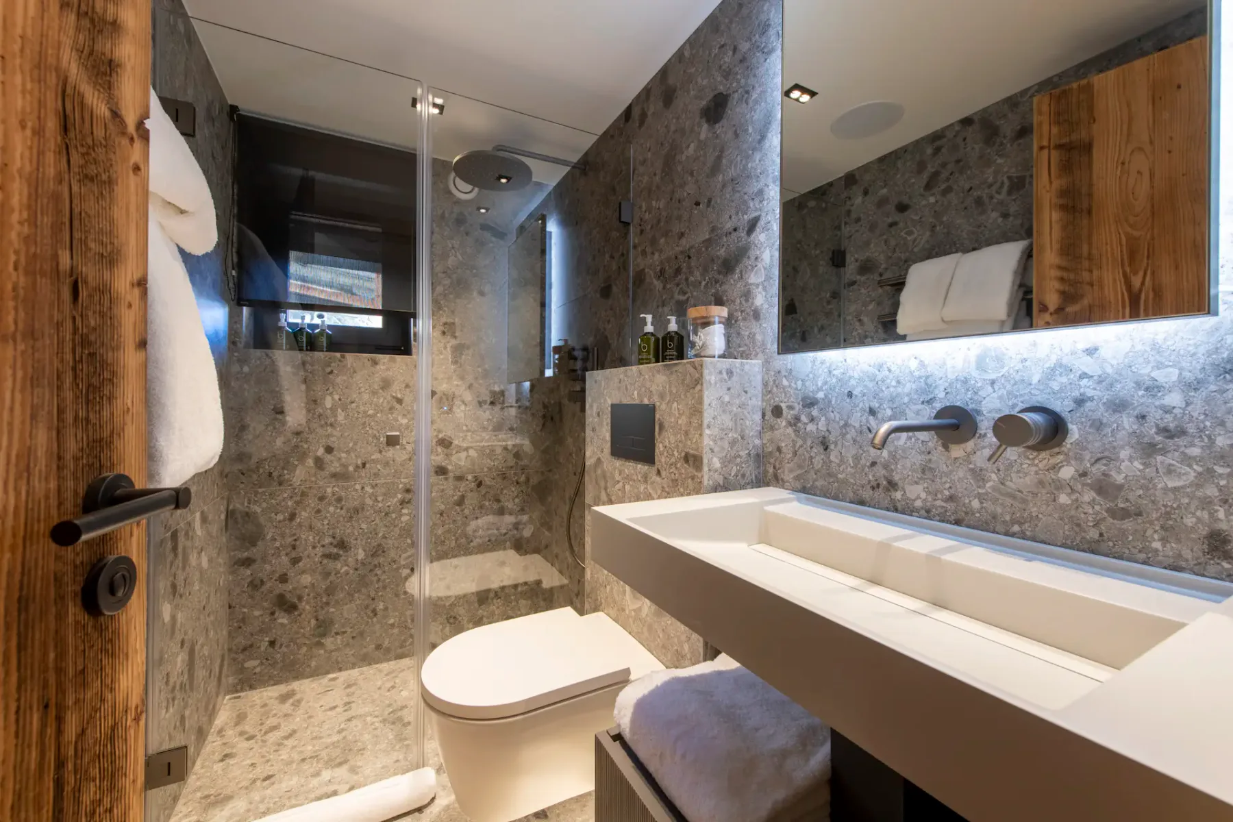 Modern bathroom with stone tile walls, a glass-enclosed shower, wall-mounted toilet, rectangular sink with sleek faucet, and a wooden door. Towels and toiletries are neatly arranged.