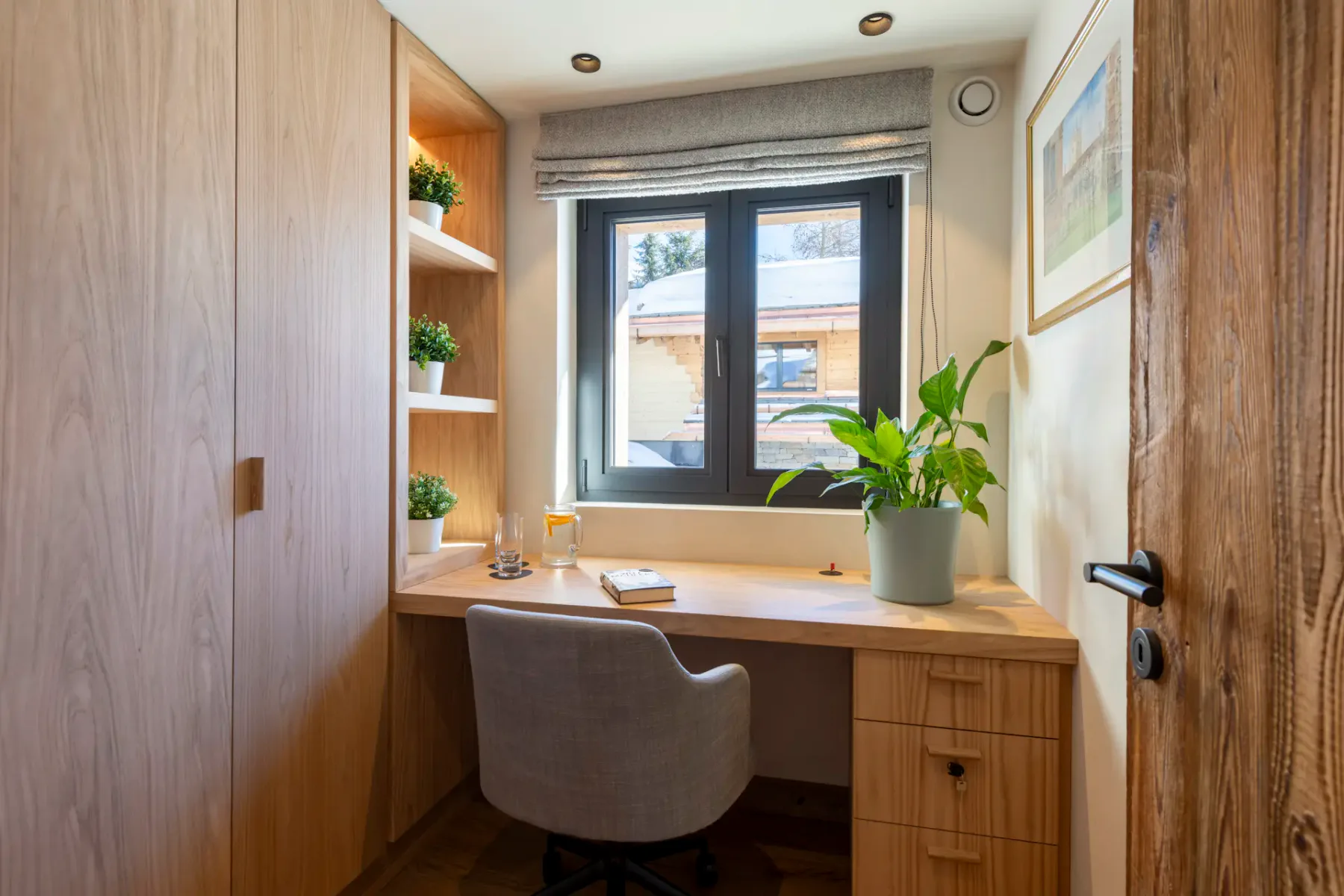 Compact home office with wooden desk, gray chair, shelves with plants, window with a blind, and a potted plant on the desk.