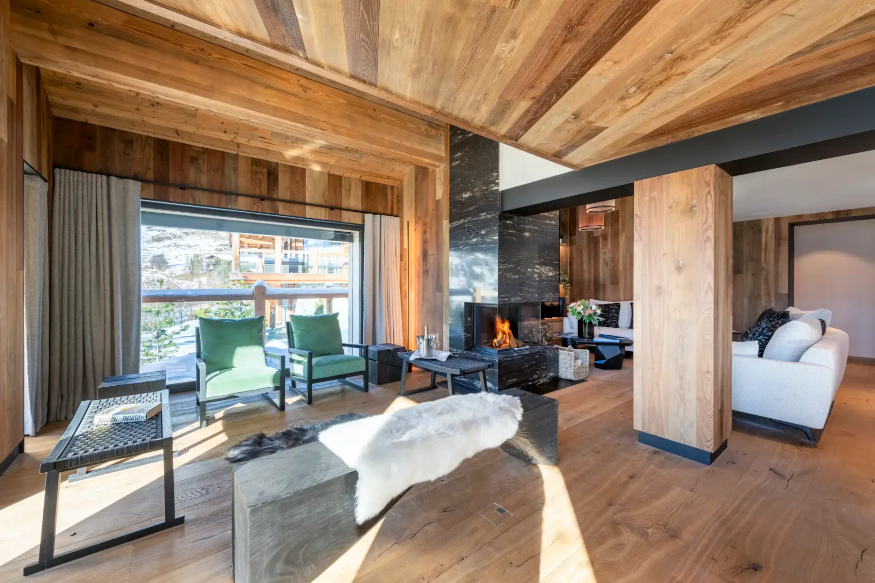 Modern living room with wooden walls and ceiling, featuring a central black fireplace, cozy sofas with dark pillows, a wall-mounted TV, and large windows with snowy mountain views.