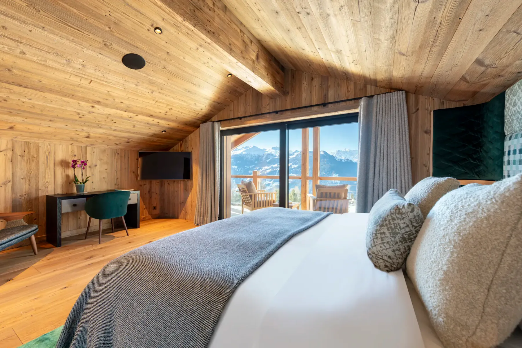 A cozy bedroom with wooden ceiling and floors, a large bed, and a balcony offering a mountain view. Two chairs are on the balcony, and light-colored curtains frame the sliding doors.