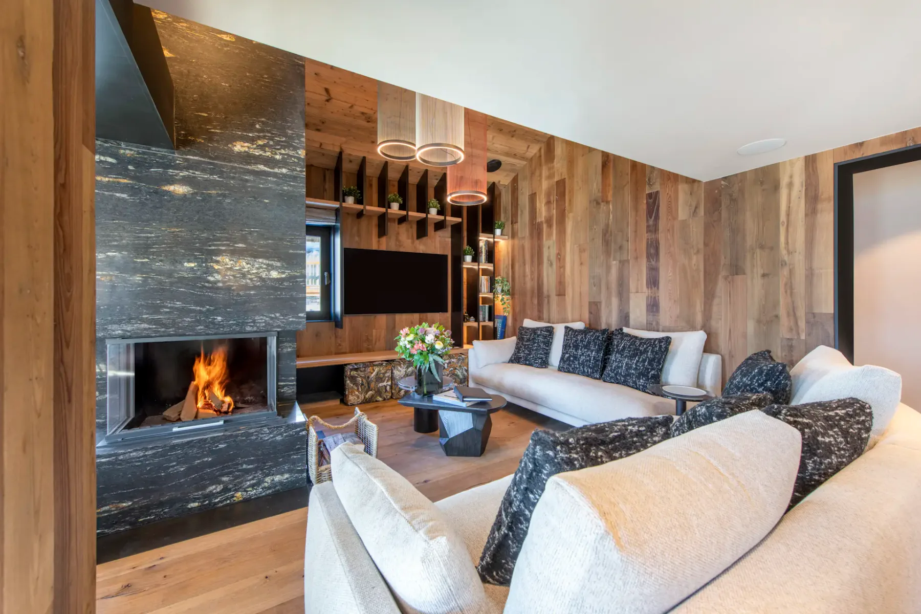 Modern living room with wooden walls and ceiling, featuring a central black fireplace, cozy sofas with dark pillows, a wall-mounted TV, and large windows with snowy mountain views.