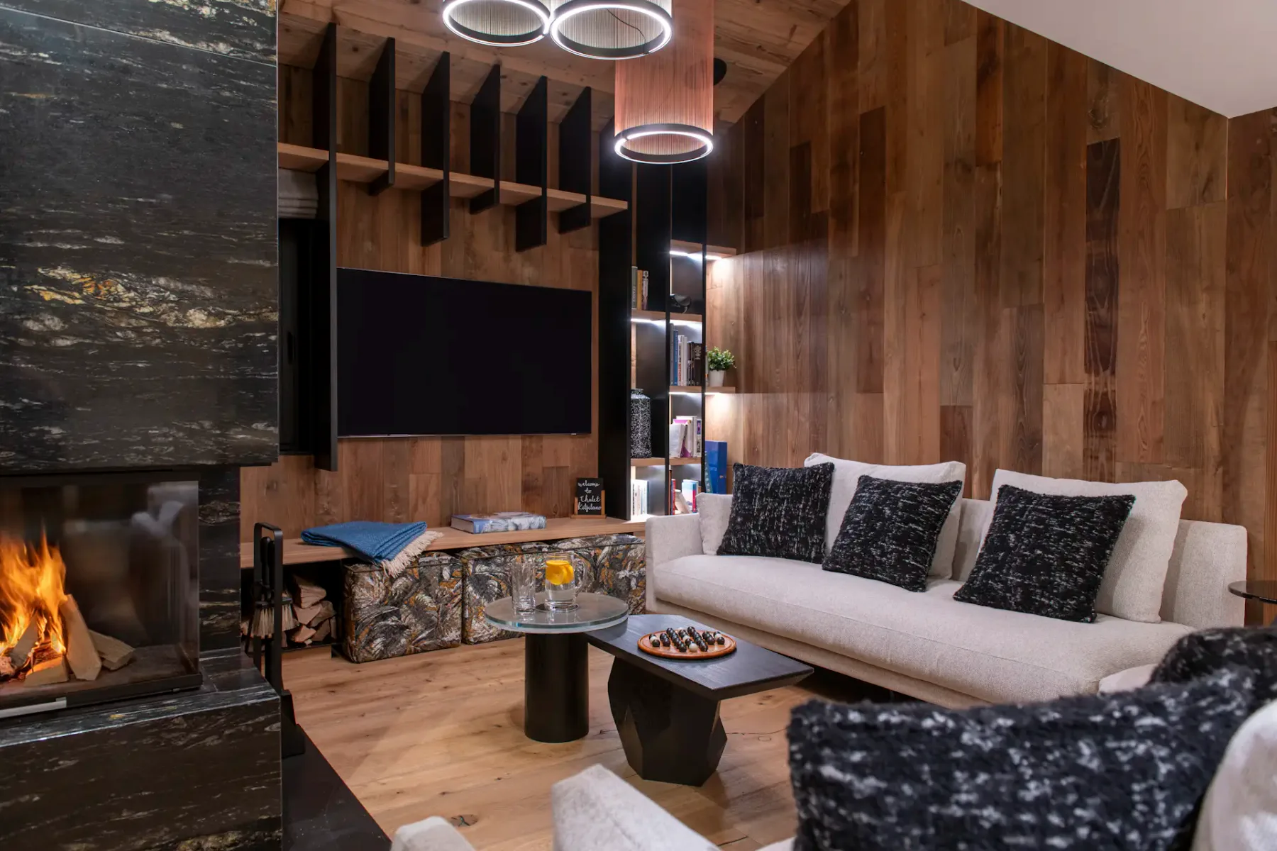 Modern living room with wooden walls and ceiling, featuring a central black fireplace, cozy sofas with dark pillows, a wall-mounted TV, and large windows with snowy mountain views.