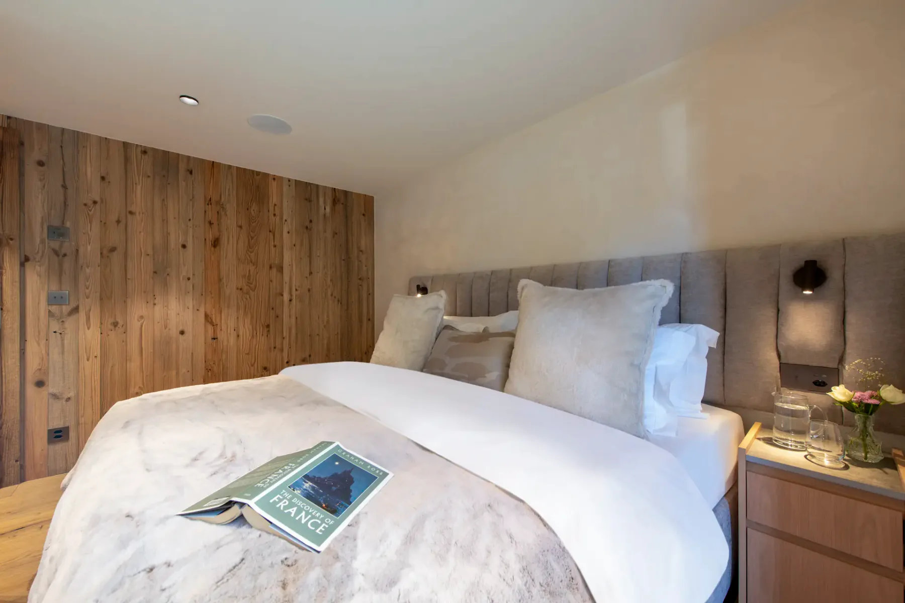 A cozy bedroom with a wooden accent wall, plush bedding, wall-mounted lamps, and a book titled