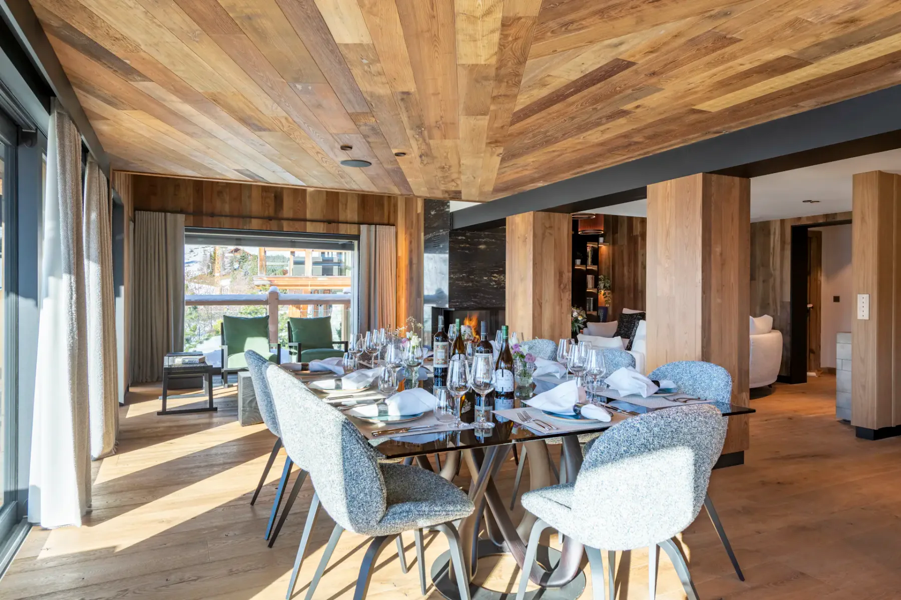 Modern dining area with a wooden ceiling, large windows showcasing a mountain view. A set table with chairs is in the foreground and a kitchen is visible in the background.
