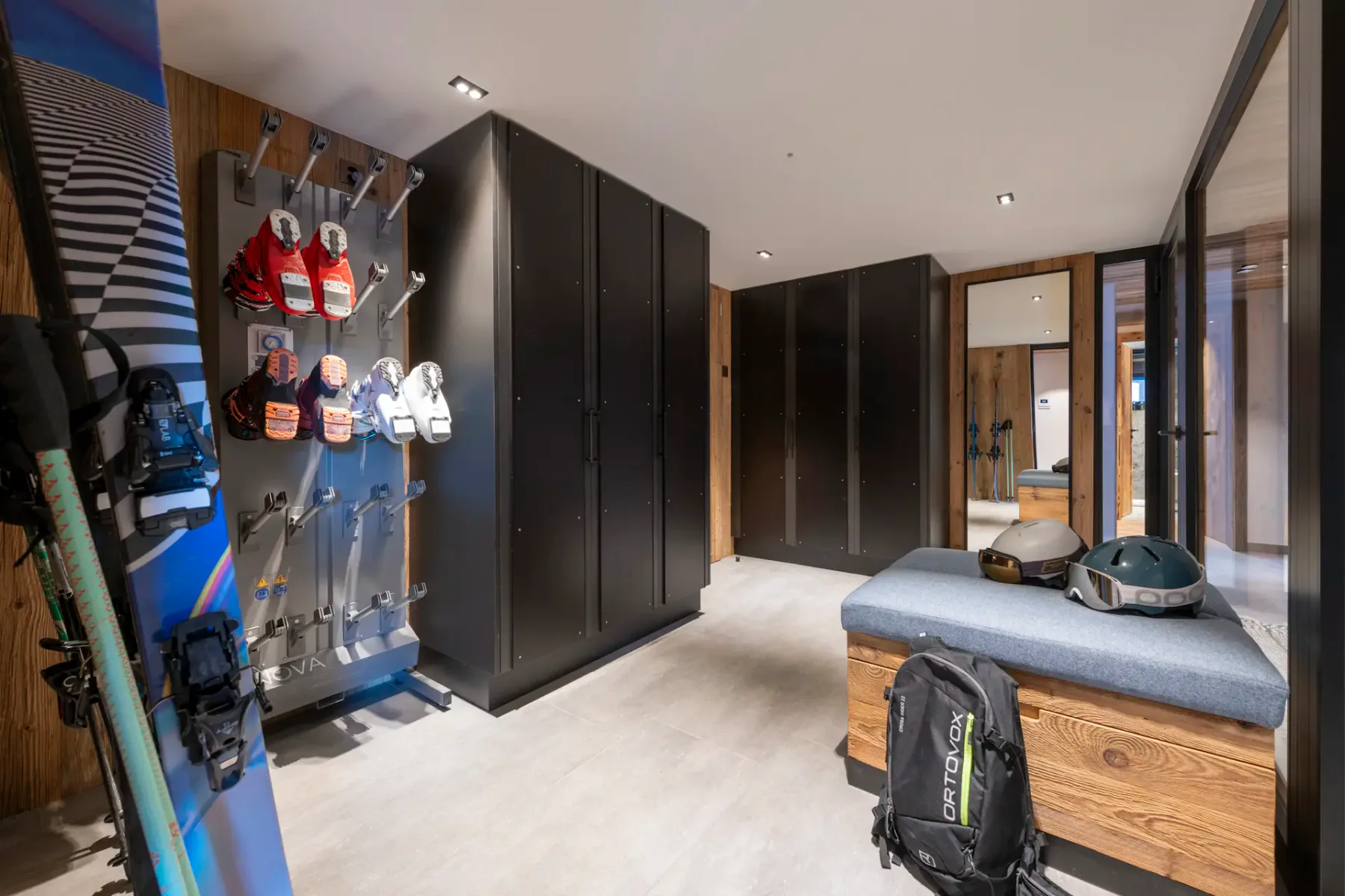 Ski room with storage lockers, ski racks with mounted boots, a bench with helmets and a backpack, and a snowboard on the left.