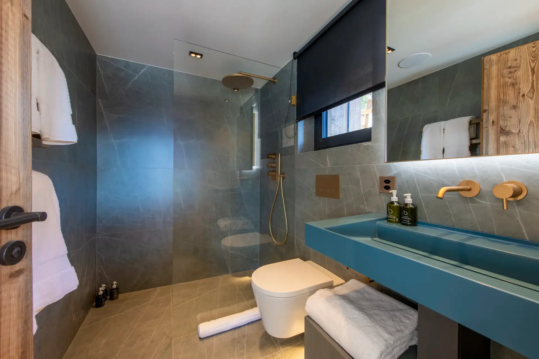 Modern bathroom with dark tiled walls, a walk-in shower, blue sink, and round toilet. Towels are neatly folded. Black roller blind covers a window above the sink.