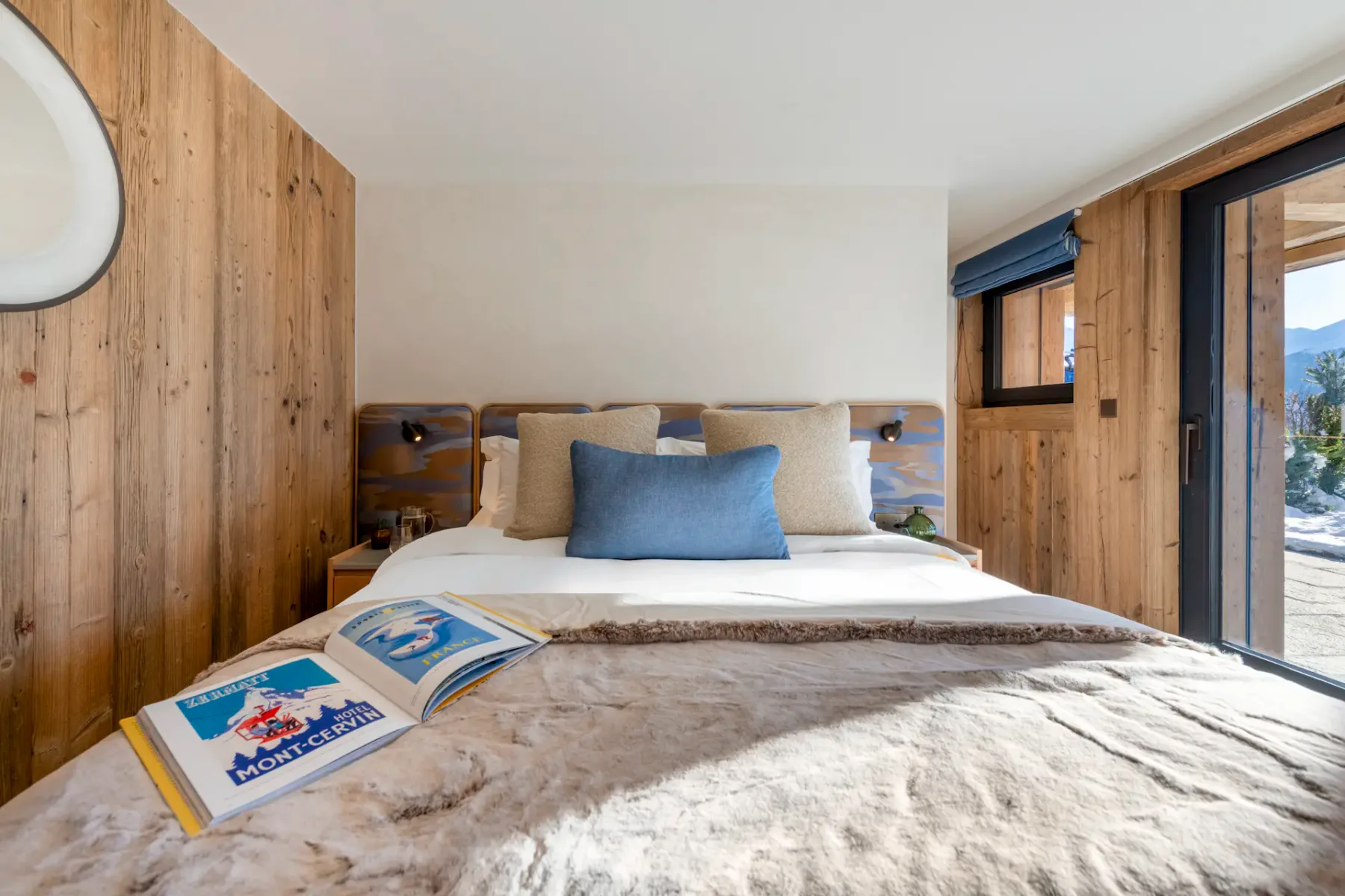 A bright bedroom with a double bed, beige and blue decor, bedside tables, and a scenic mountain view through large sliding glass doors. An open magazine rests on the bed.
