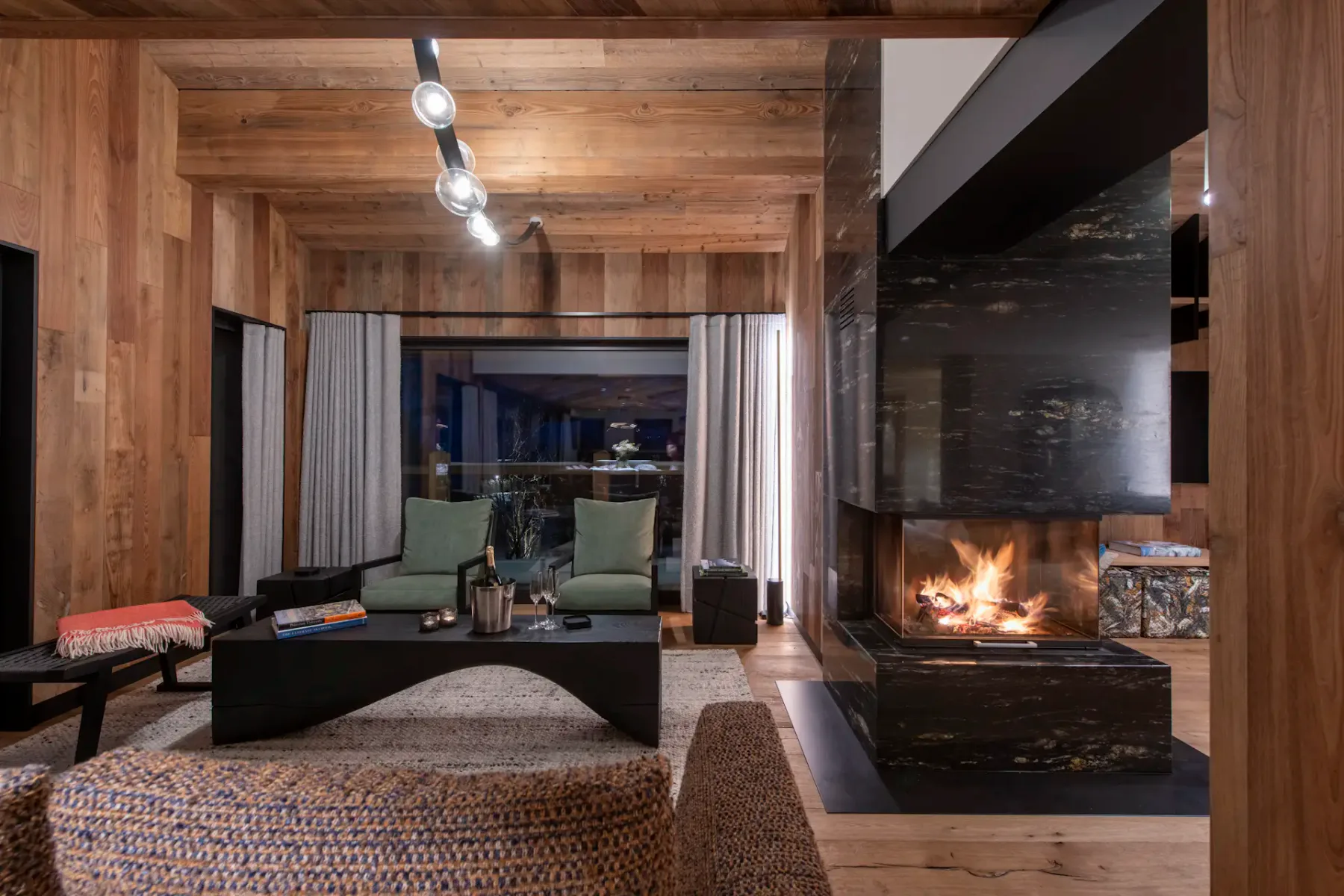 Modern living room with wooden walls and ceiling, featuring a central black fireplace, cozy sofas with dark pillows, a wall-mounted TV, and large windows with snowy mountain views.