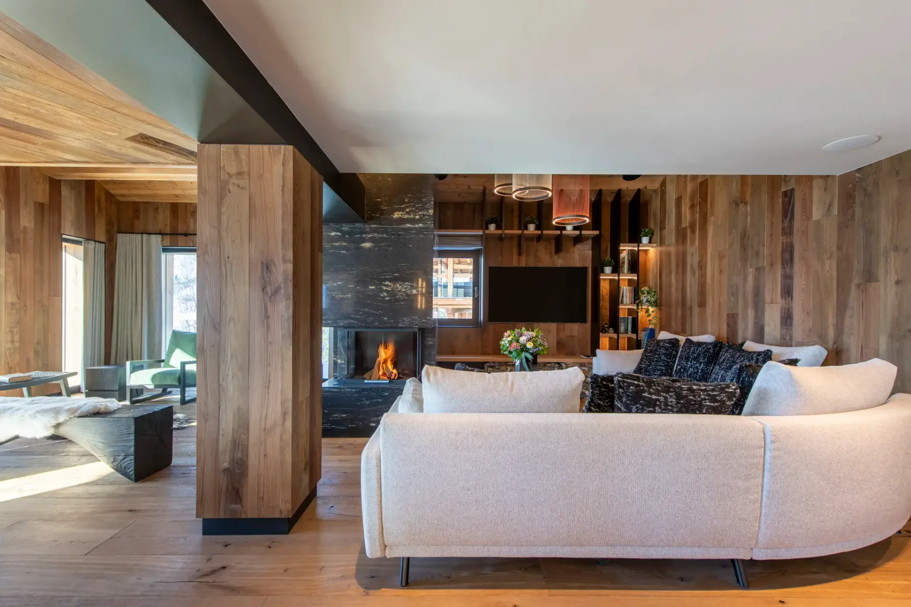 Modern living room with wooden walls and ceiling, featuring a central black fireplace, cozy sofas with dark pillows, a wall-mounted TV, and large windows with snowy mountain views.