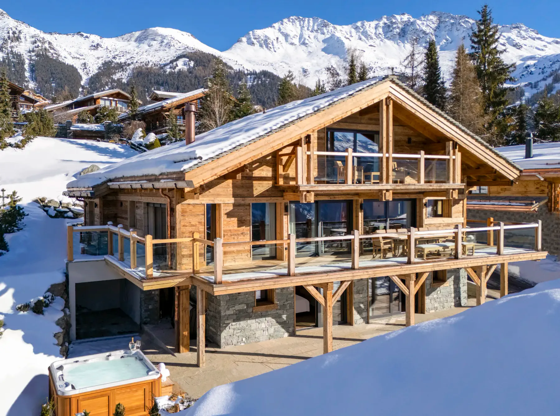 A wooden chalet with large windows and balconies, situated in a snowy mountainous area with clear blue skies. Nearby are trees and other chalets.