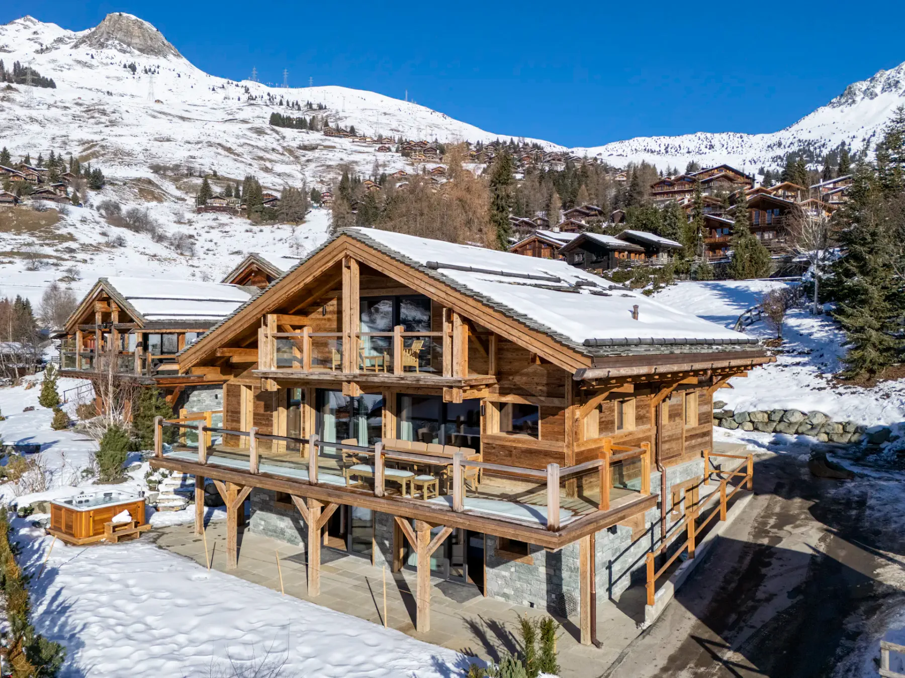 A wooden chalet with large windows and balconies, situated in a snowy mountainous area with clear blue skies. Nearby are trees and other chalets.
