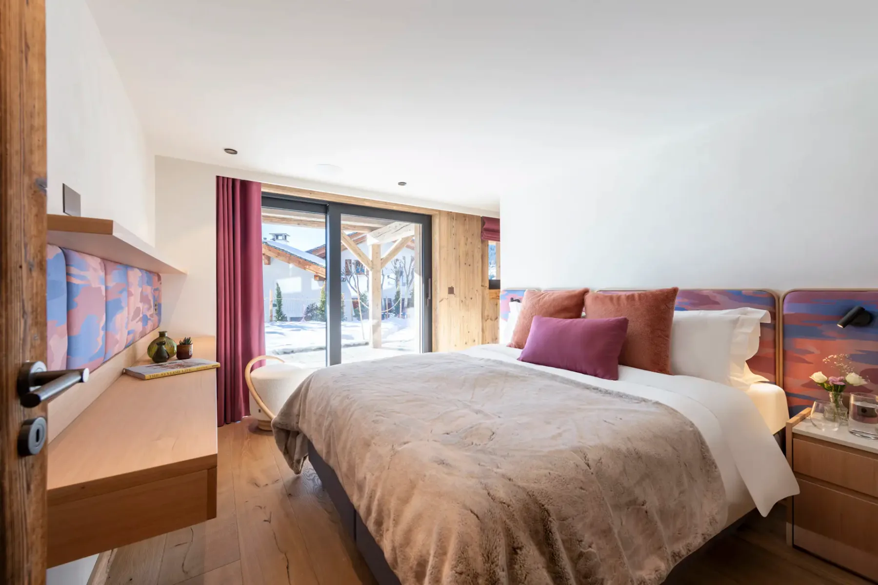 Modern bedroom with a large bed, wooden floors, sliding glass doors, purple curtains, and outdoor view. Walls are white with a wooden door, and a floating shelf on the left.