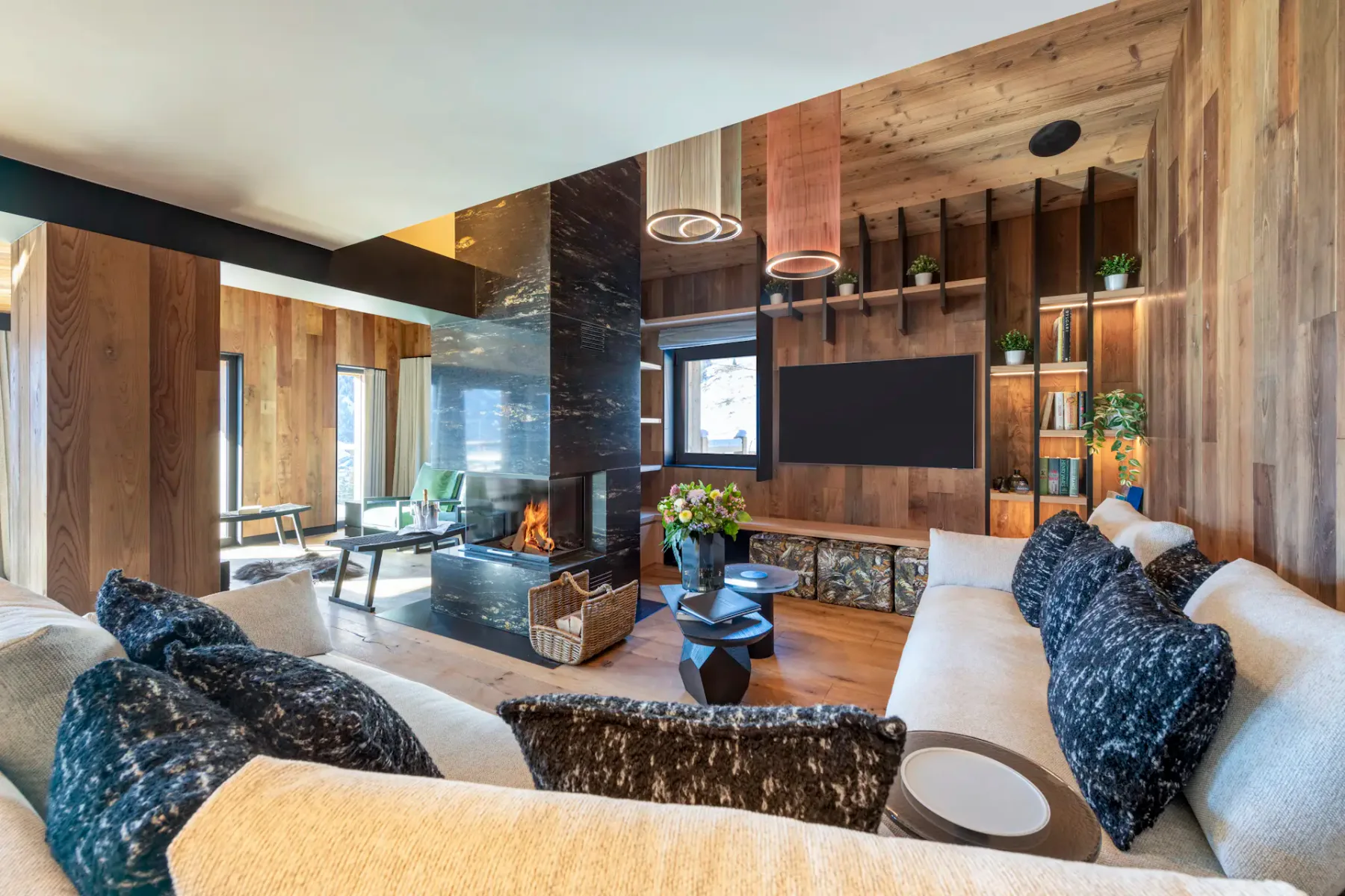 Modern living room with wooden walls and ceiling, featuring a central black fireplace, cozy sofas with dark pillows, a wall-mounted TV, and large windows with snowy mountain views.