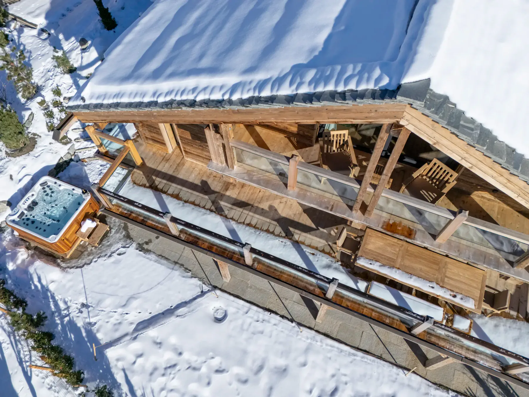 A wooden chalet with large windows and balconies, situated in a snowy mountainous area with clear blue skies. Nearby are trees and other chalets.