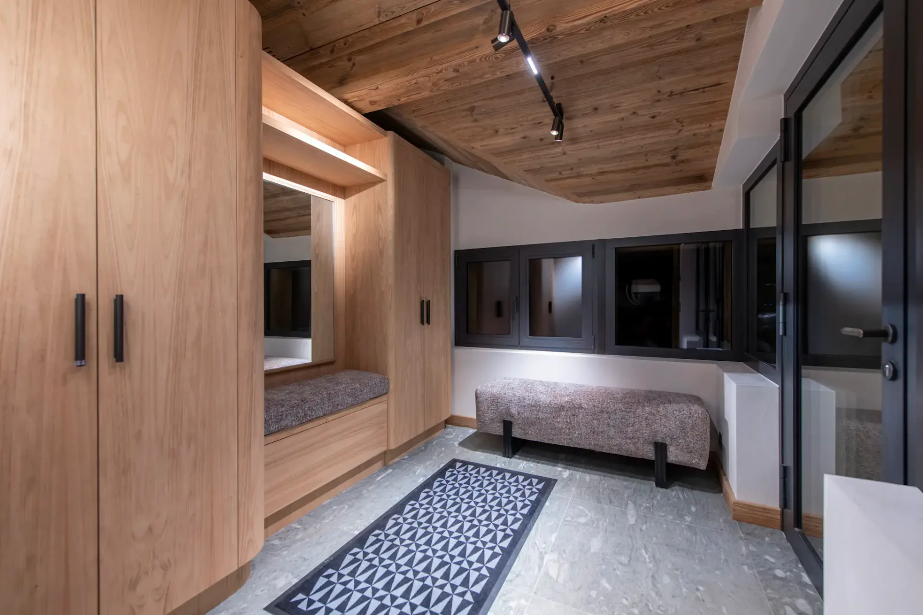 A modern entryway with wooden ceiling, closet, built-in seating, patterned rug, and large windows with black frames.