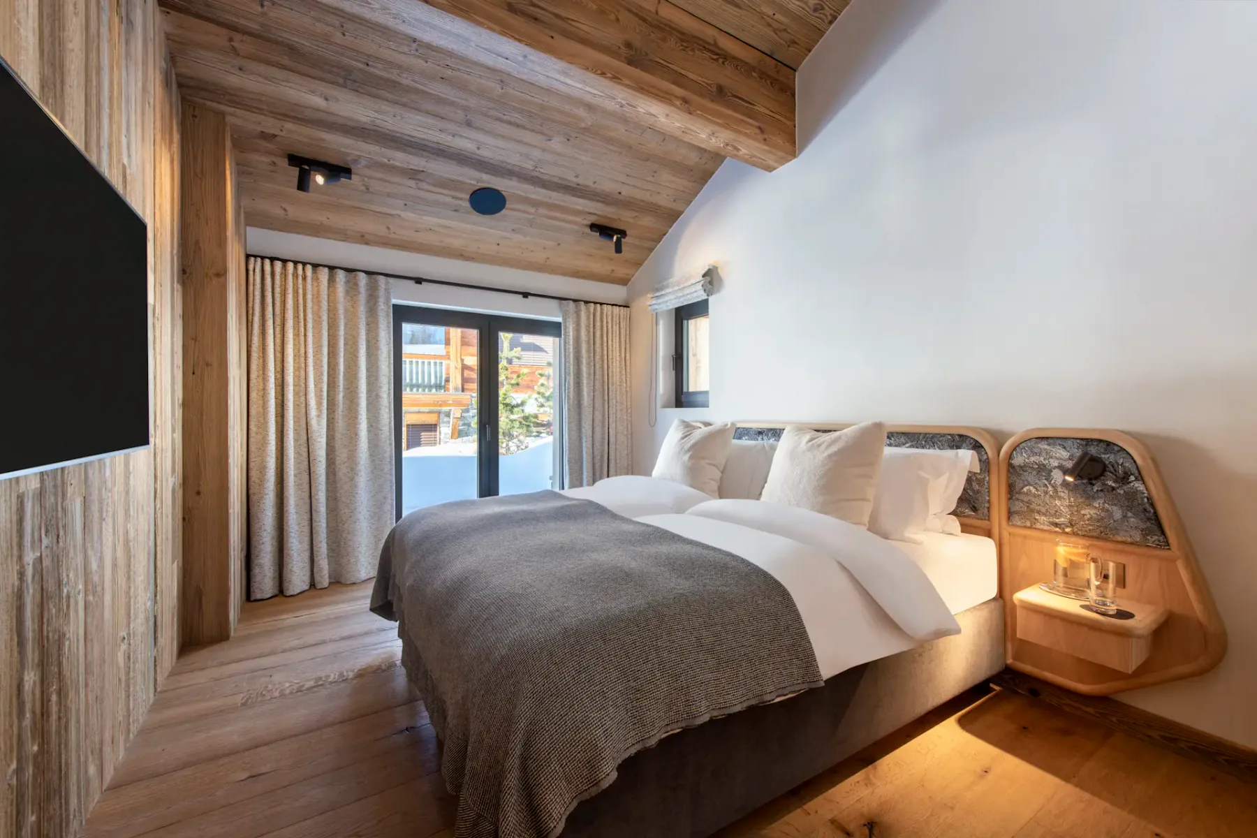 Modern bedroom with wooden ceiling and floor, featuring a double bed with white linens, gray throw, and two pillows. There's a large window with curtains and a wall-mounted flat-screen TV.